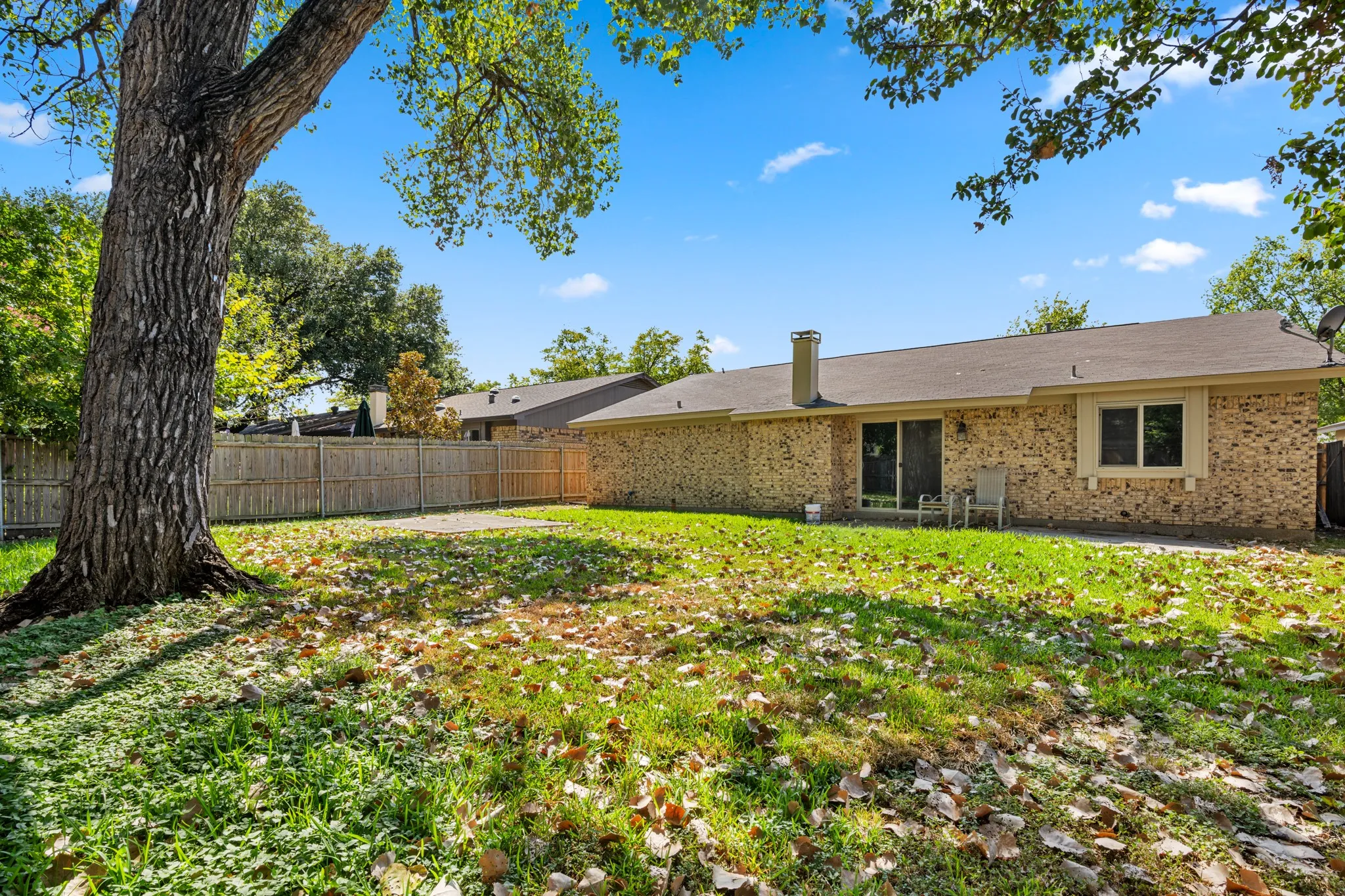 Spacious private backyard featuring a large mature shade tree, expansive grassy area, and wood privacy fencing. A concrete patio provides a great space for outdoor seating and entertaining, with direct access to the home through the sliding glass door. The generous yard offers plenty of room for outdoor enjoyment, gardening, or play.