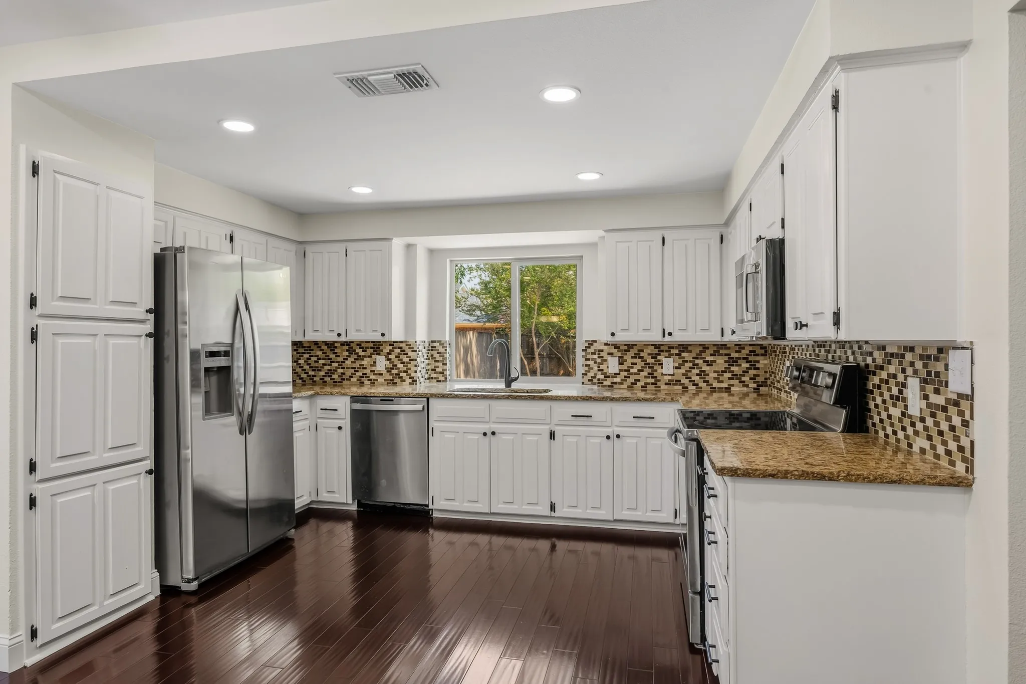 Bright and inviting kitchen featuring an abundance of crisp white cabinetry, granite countertops, and a stylish mosaic tile backsplash. Stainless steel appliances, including a side-by-side refrigerator and dishwasher, complement the space, while a large window above the sink provides natural light and a pleasant view. Rich wood flooring adds warmth and contrast to this well-appointed kitchen.