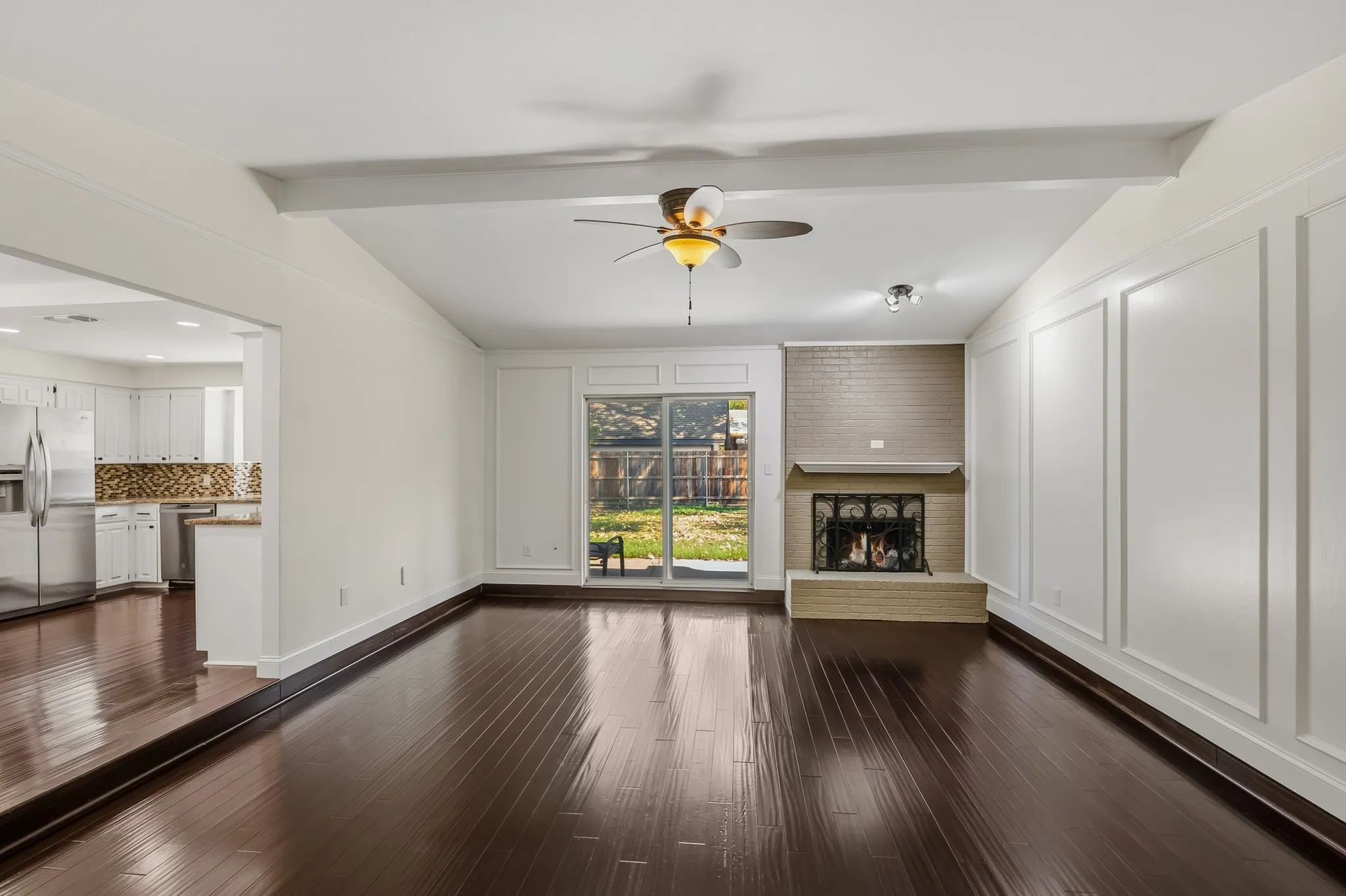 Spacious and inviting living room featuring rich wood-look flooring, crisp neutral walls, and elegant decorative wall paneling. A classic brick fireplace with raised hearth creates a warm focal point, while the large glass patio door fills the room with natural light and provides views of the backyard. The open layout flows seamlessly into the kitchen, creating a comfortable space for everyday living and entertaining.