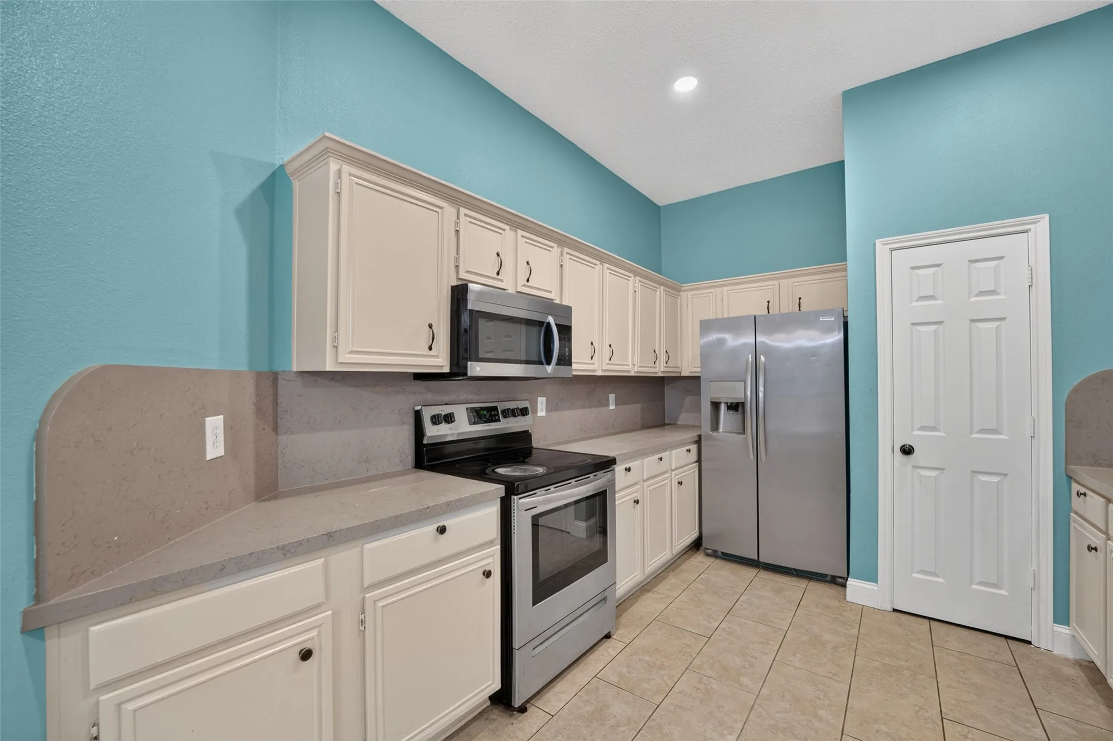 Kitchen featuring vaulted ceilings, light-toned cabinetry with dark hardware, stainless steel appliances, solid surface countertops, and tile flooring