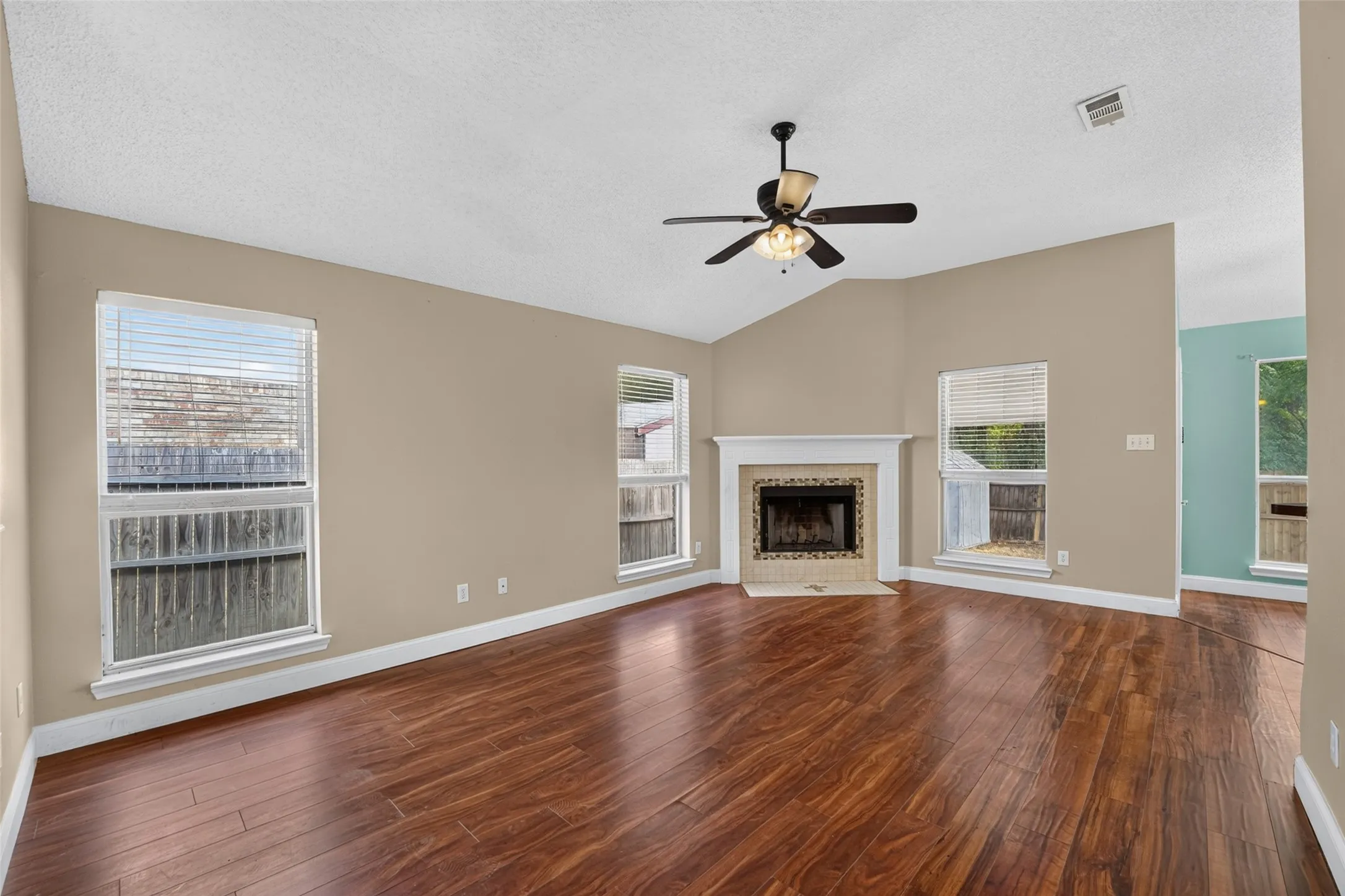 Vaulted ceiling with a ceiling fan, wood-finish flooring, fireplace with a tile surround and white mantel, and multiple windows