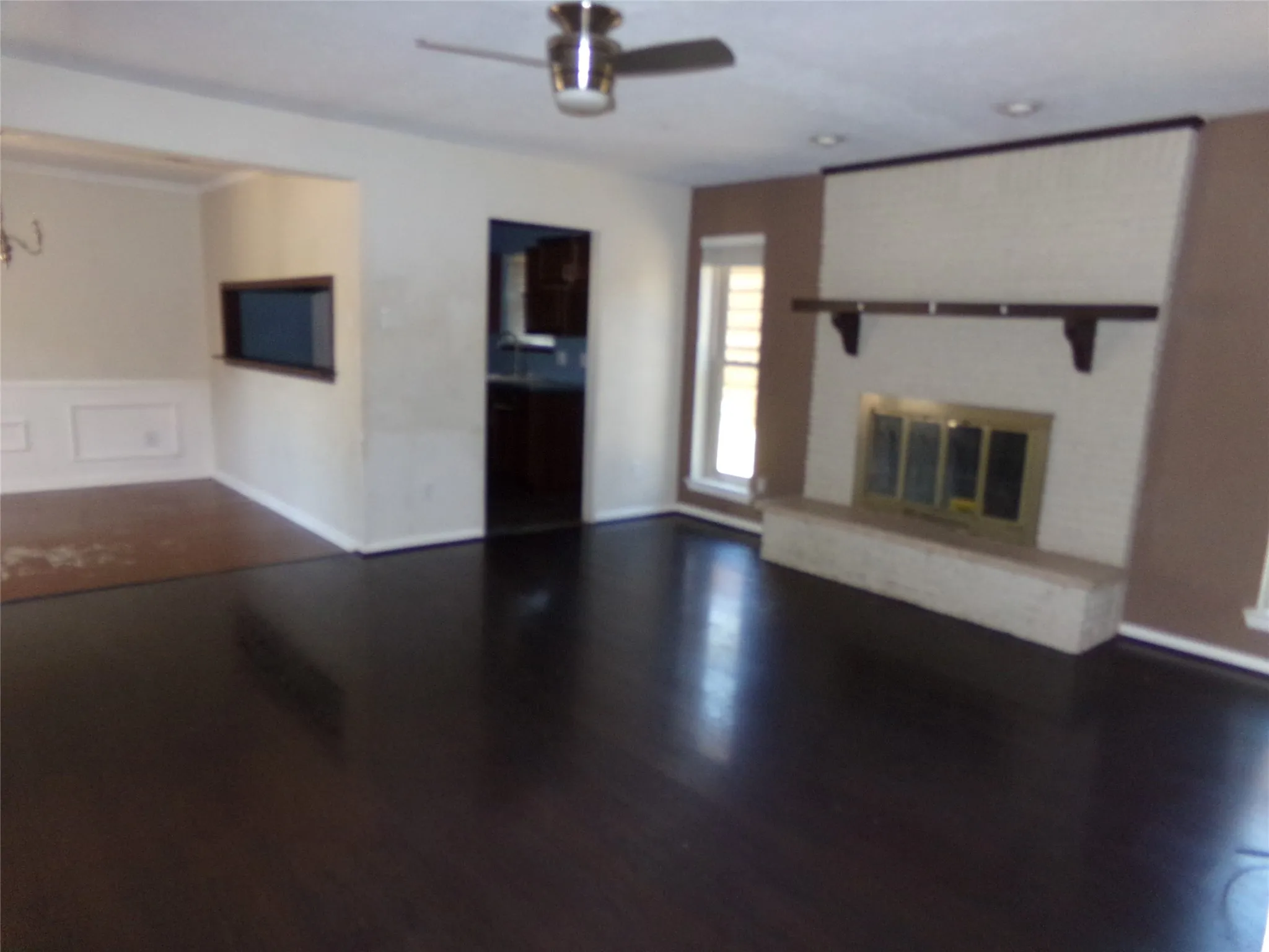 Dark wood-finish flooring, white brick fireplace with a hearth, pass-through window, and a ceiling fan