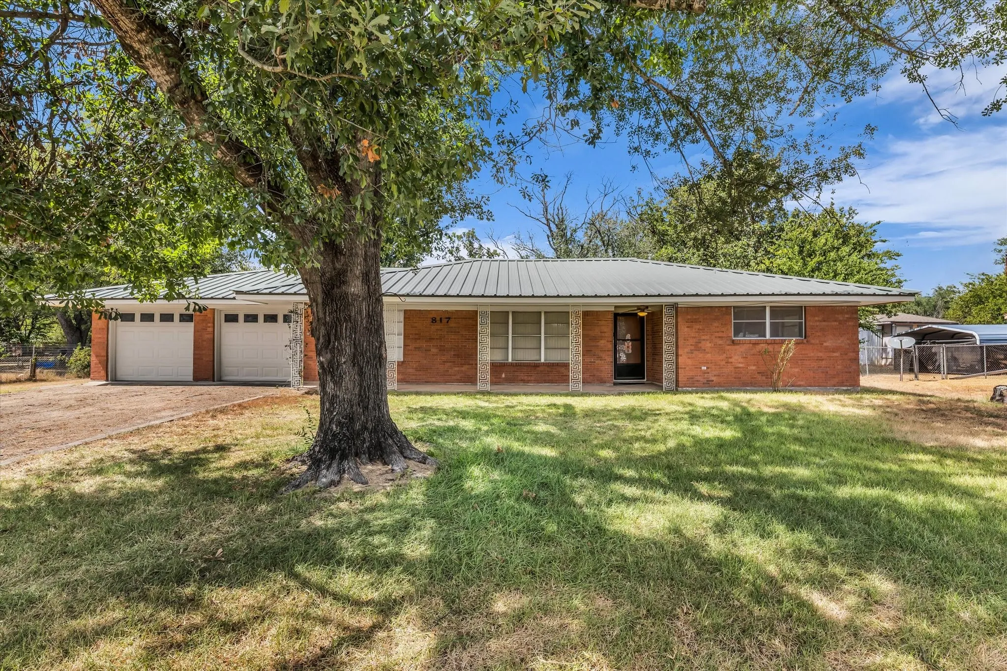 Street View of 817 Holly Lane, featuring a brick exterior, metal roof, 2 car garage, large covered porch, and paved driveway.