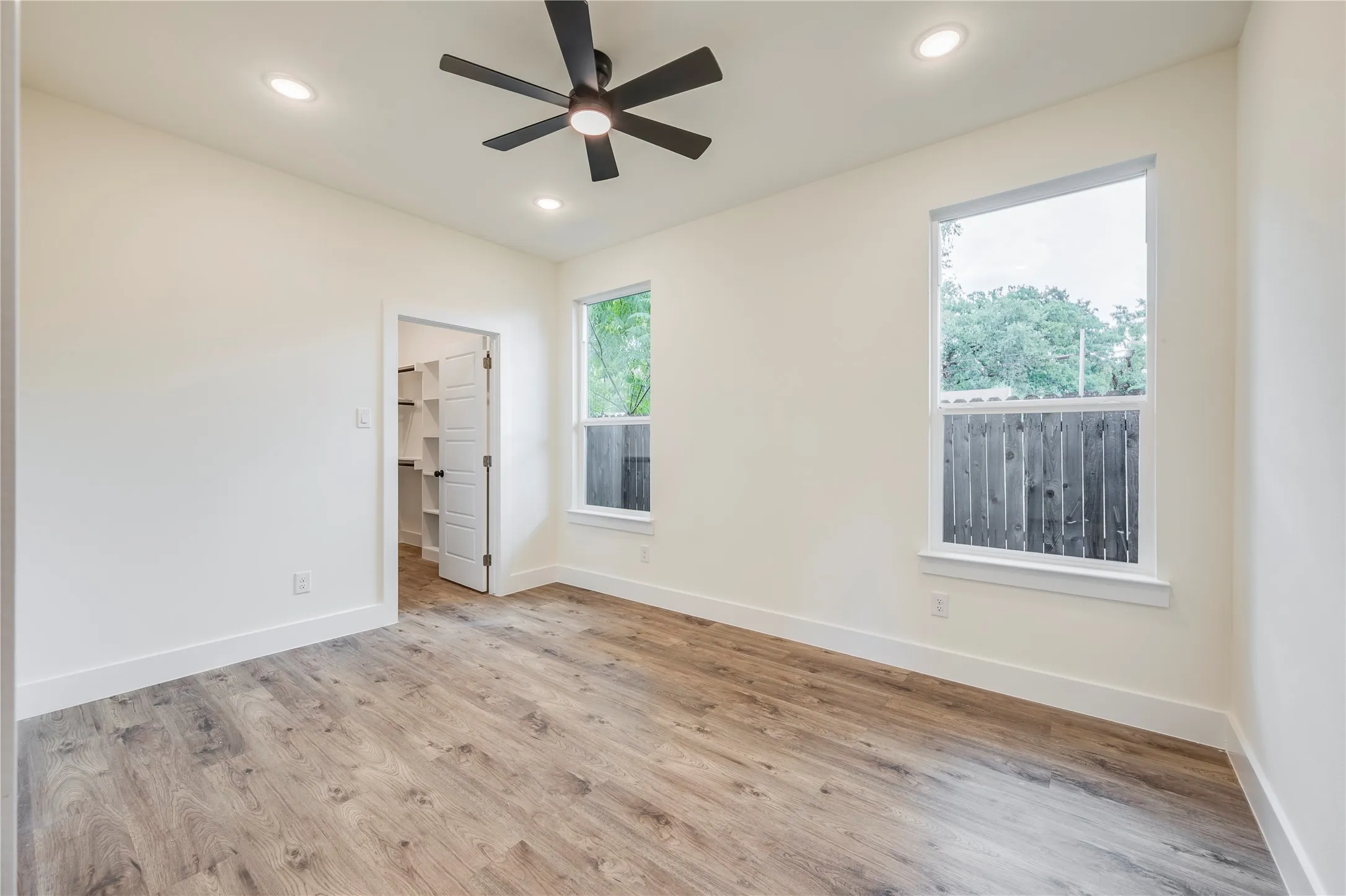 Spacious room with wood-finish flooring, two large windows, a ceiling fan, recessed lighting, and a walk-in closet with built-in shelving