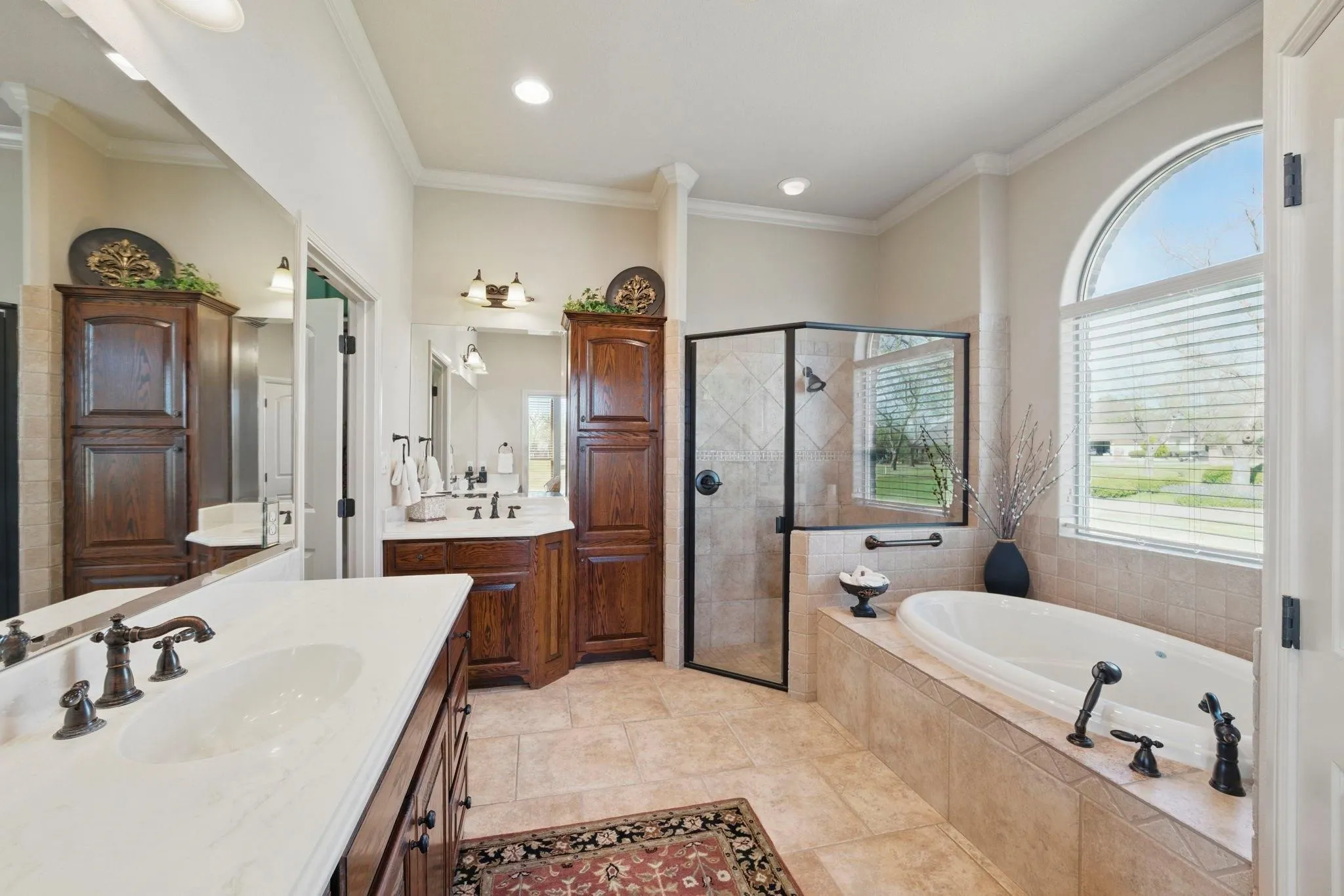 Dual vanity with white countertops, tiled flooring, glass-enclosed shower, built-in soaking tub, and arched window