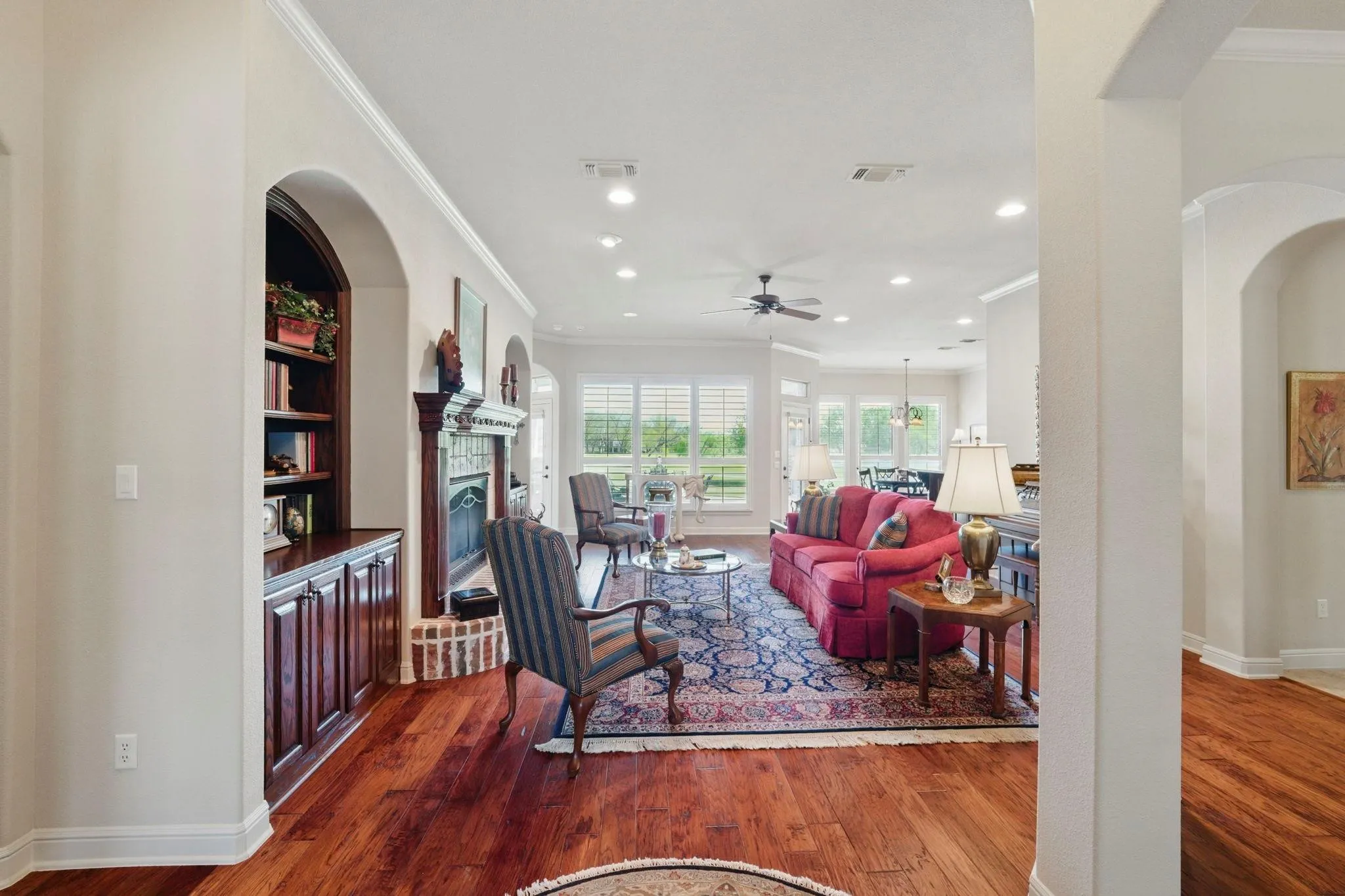 Warm wood-finish flooring throughout, featuring a built-in arched bookshelf, a fireplace with a brick surround, recessed lighting, and a ceiling fan
