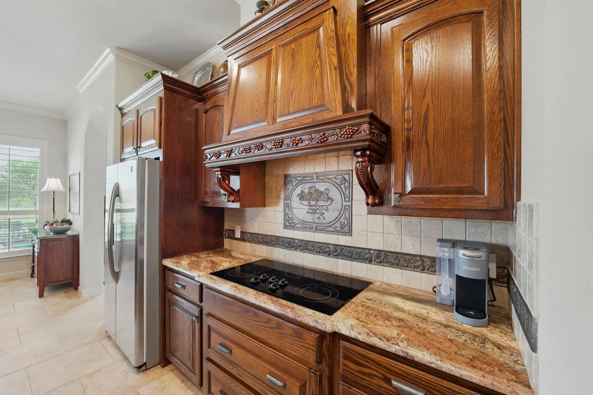 Rich wood cabinetry, patterned tile backsplash, integrated cooktop, stone countertop, and stainless steel refrigerator