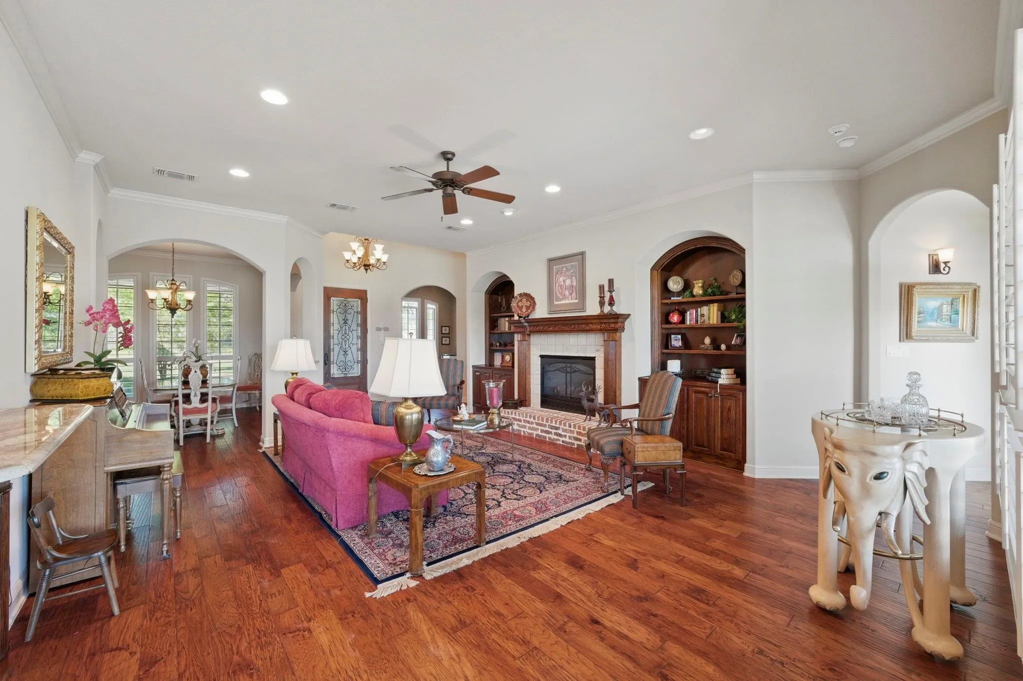 Open-concept living area featuring wood-finish flooring, a brick-surround fireplace with a wood mantel, built-in shelving, recessed lighting, and arched doorways