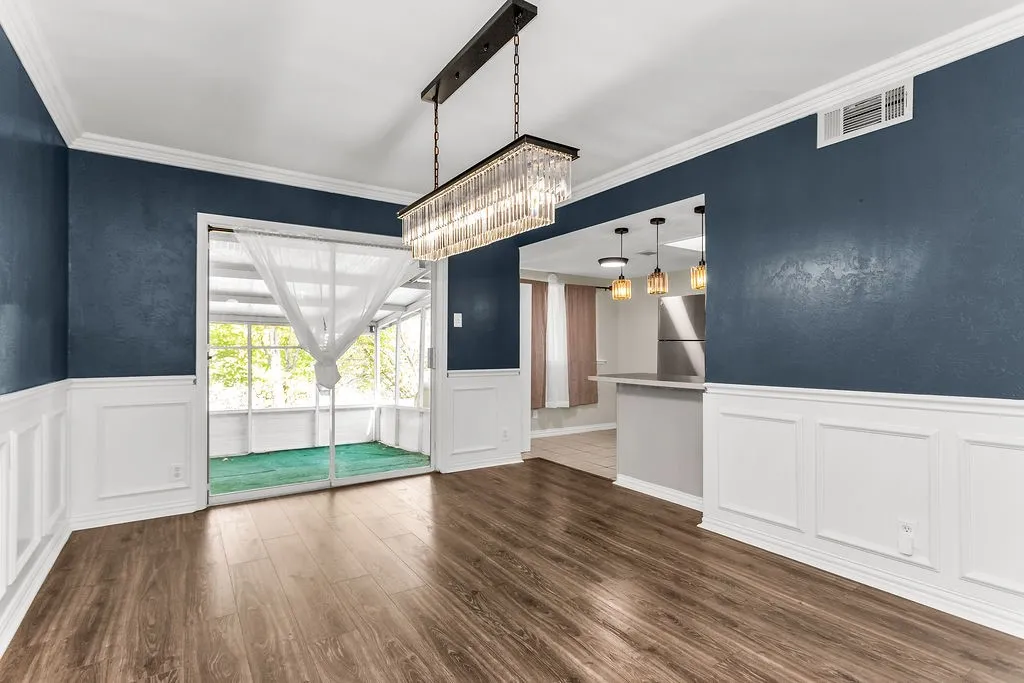 Wood-finish flooring, wainscoting, crown molding, a modern linear chandelier, and sliding glass doors to a screened enclosure