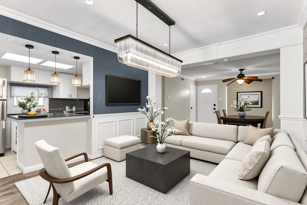 Open-concept living area featuring wood-finish flooring, a kitchen pass-through with pendant lighting, crown molding, wainscoting, and a linear chandelier