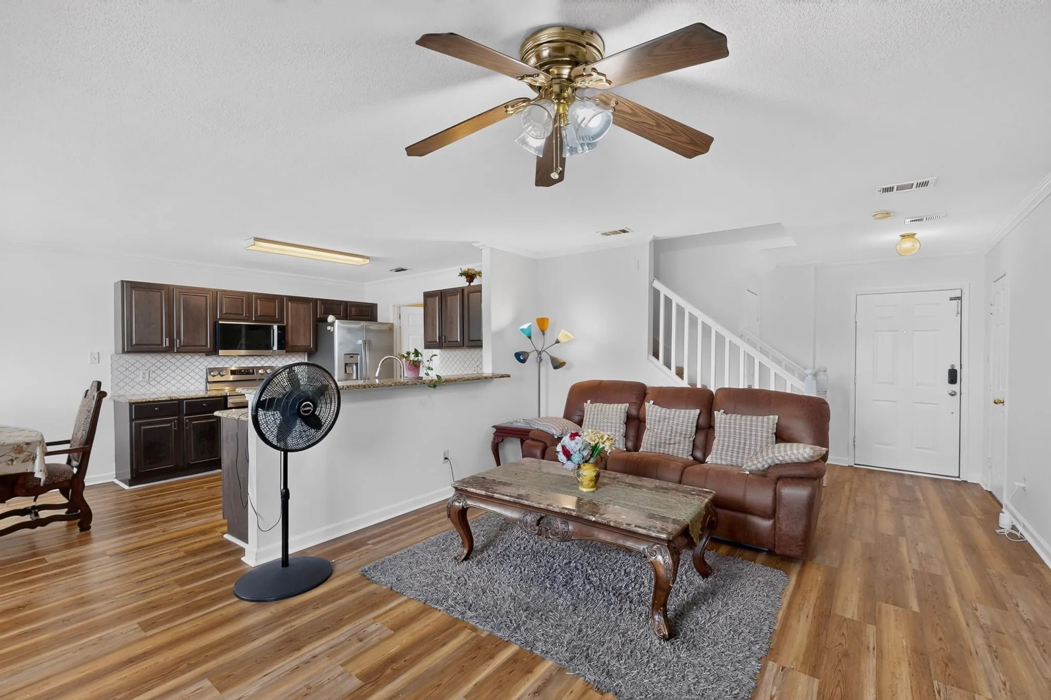 Open-concept living area featuring wood-finish flooring, a kitchen with dark wood cabinetry, stainless steel appliances, and a white subway tile backsplash, and a staircase with white railings
