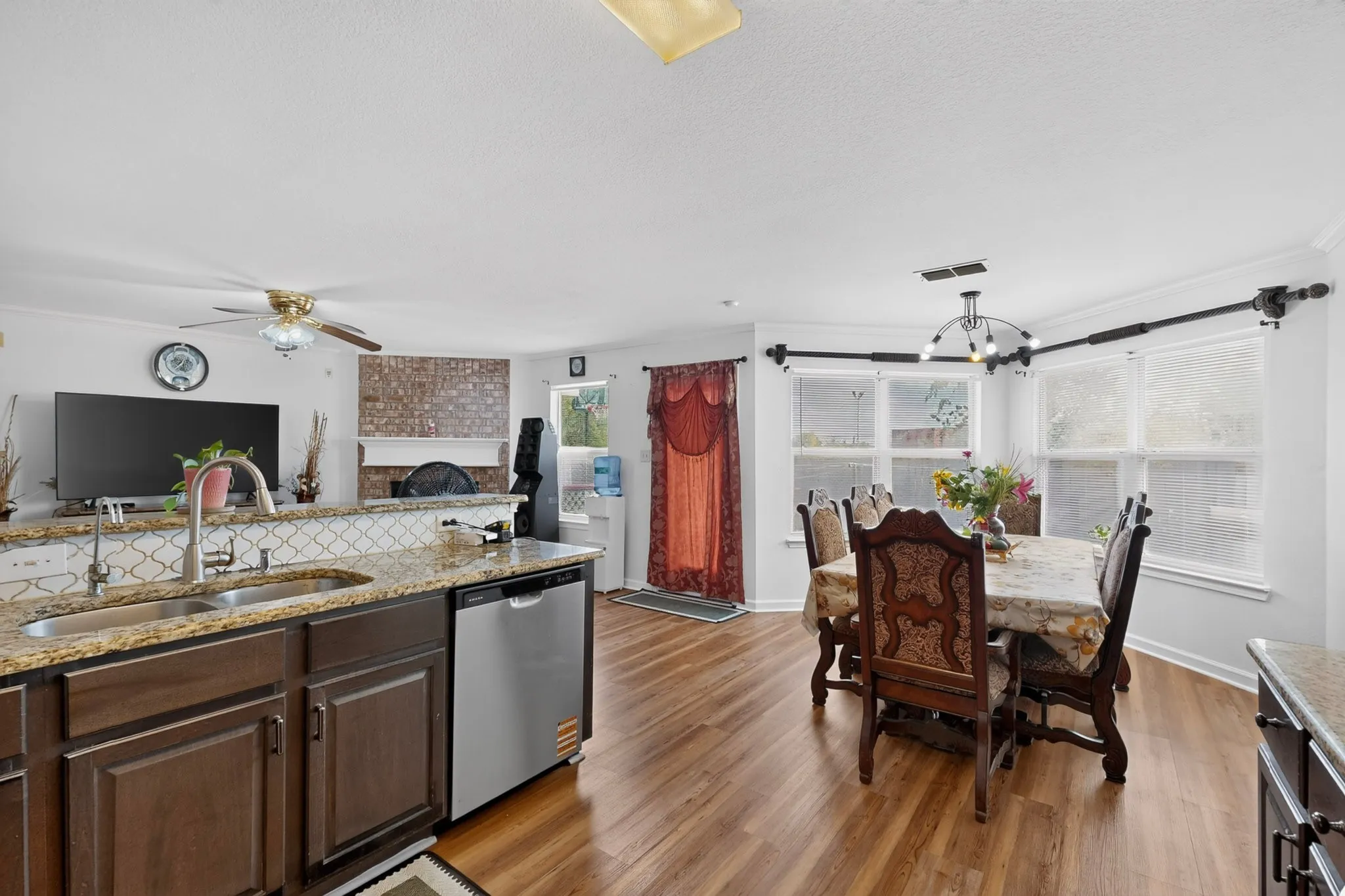 Open-concept living area featuring wood-finish flooring, a kitchen island with a double basin sink and granite countertops, and a dining area with multiple windows