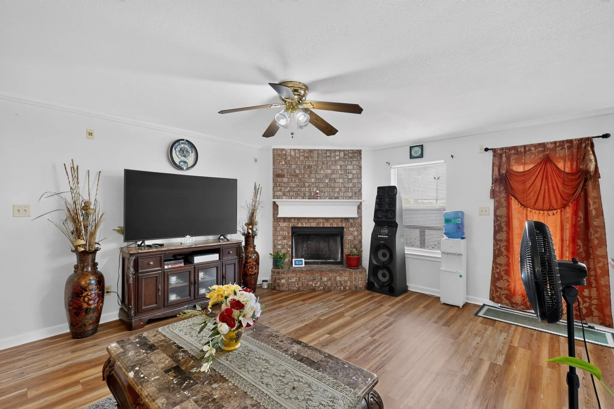 Brick fireplace with a white mantel, wood-finish flooring, white walls, ceiling fan, and white trim molding