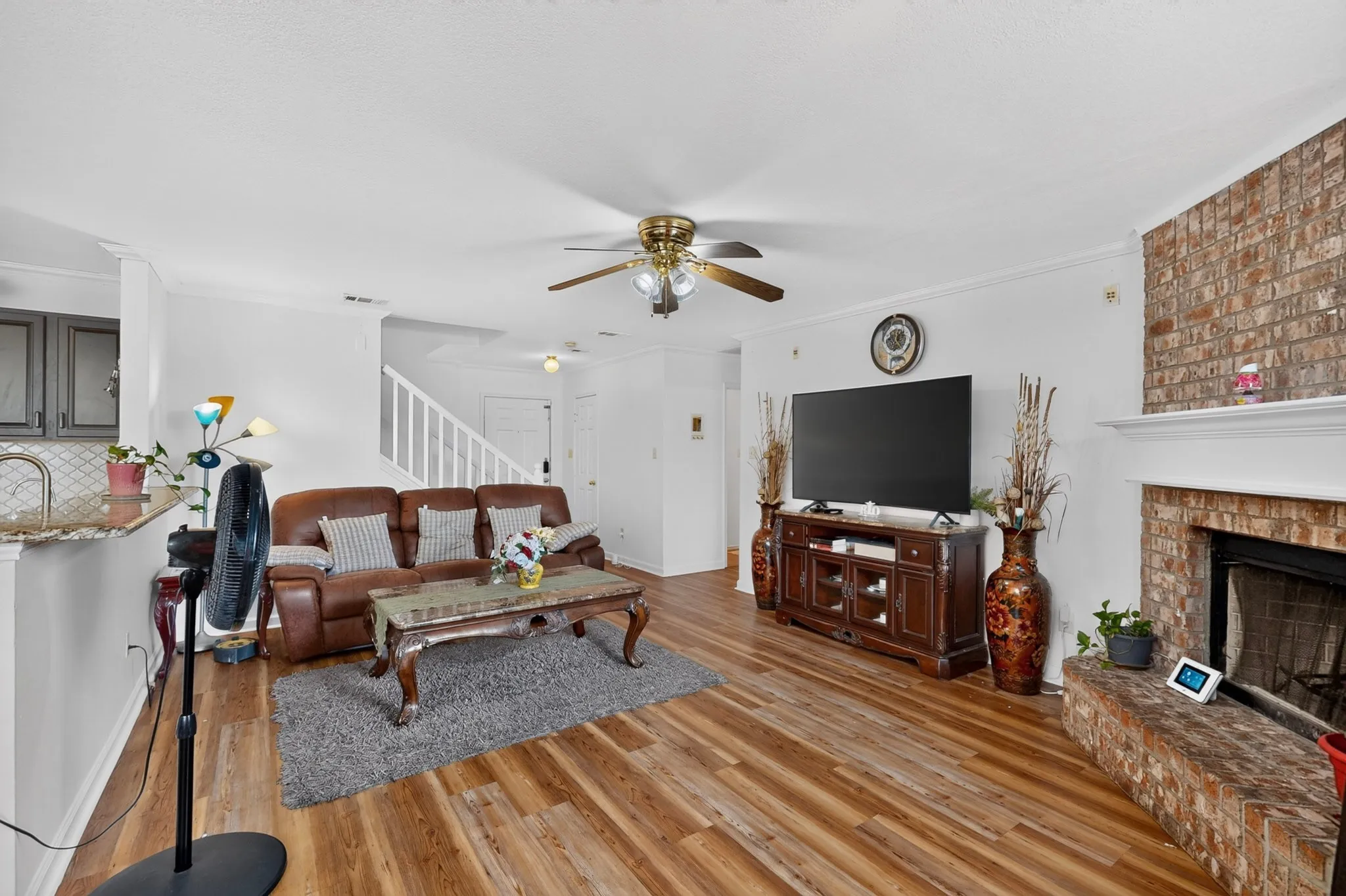 Wood-finish flooring, brick fireplace surround with white mantel, ceiling fan, and white crown molding
