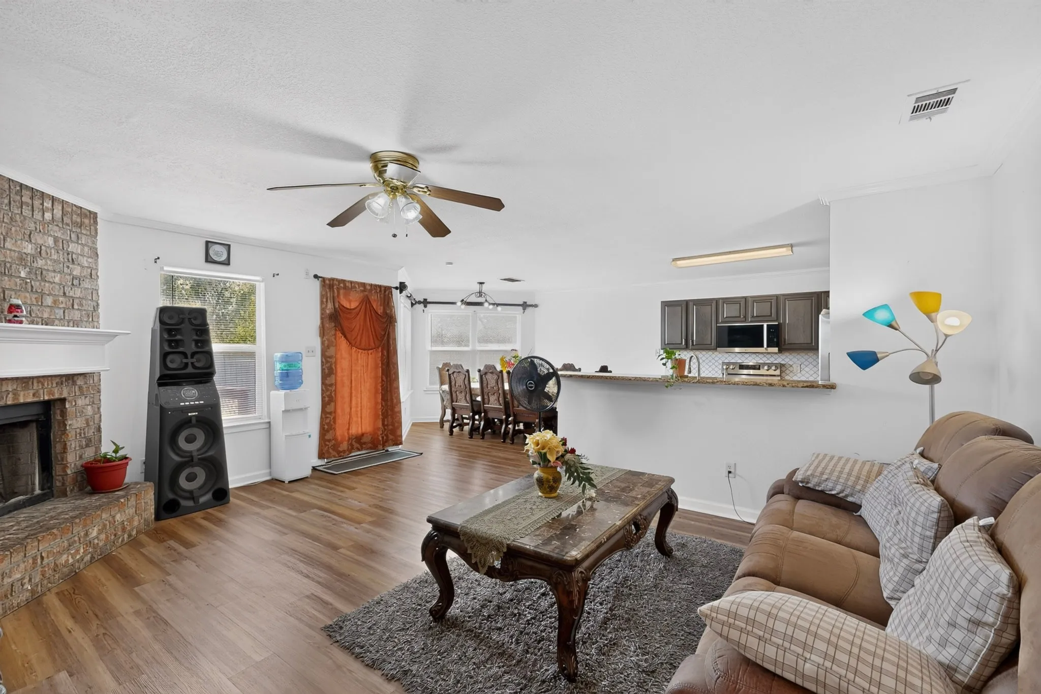 Brick-surround fireplace with a white mantle, wood-finish flooring, ceiling fan, kitchen pass-through, and dark wood-finish cabinetry