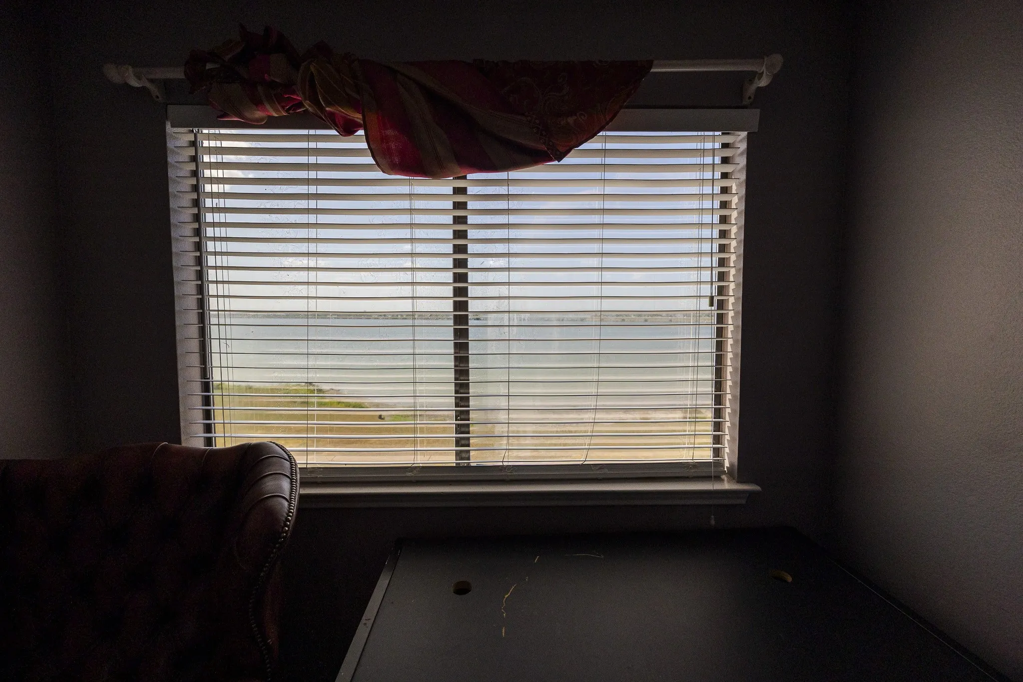 Window with horizontal blinds overlooking a body of water and distant shoreline