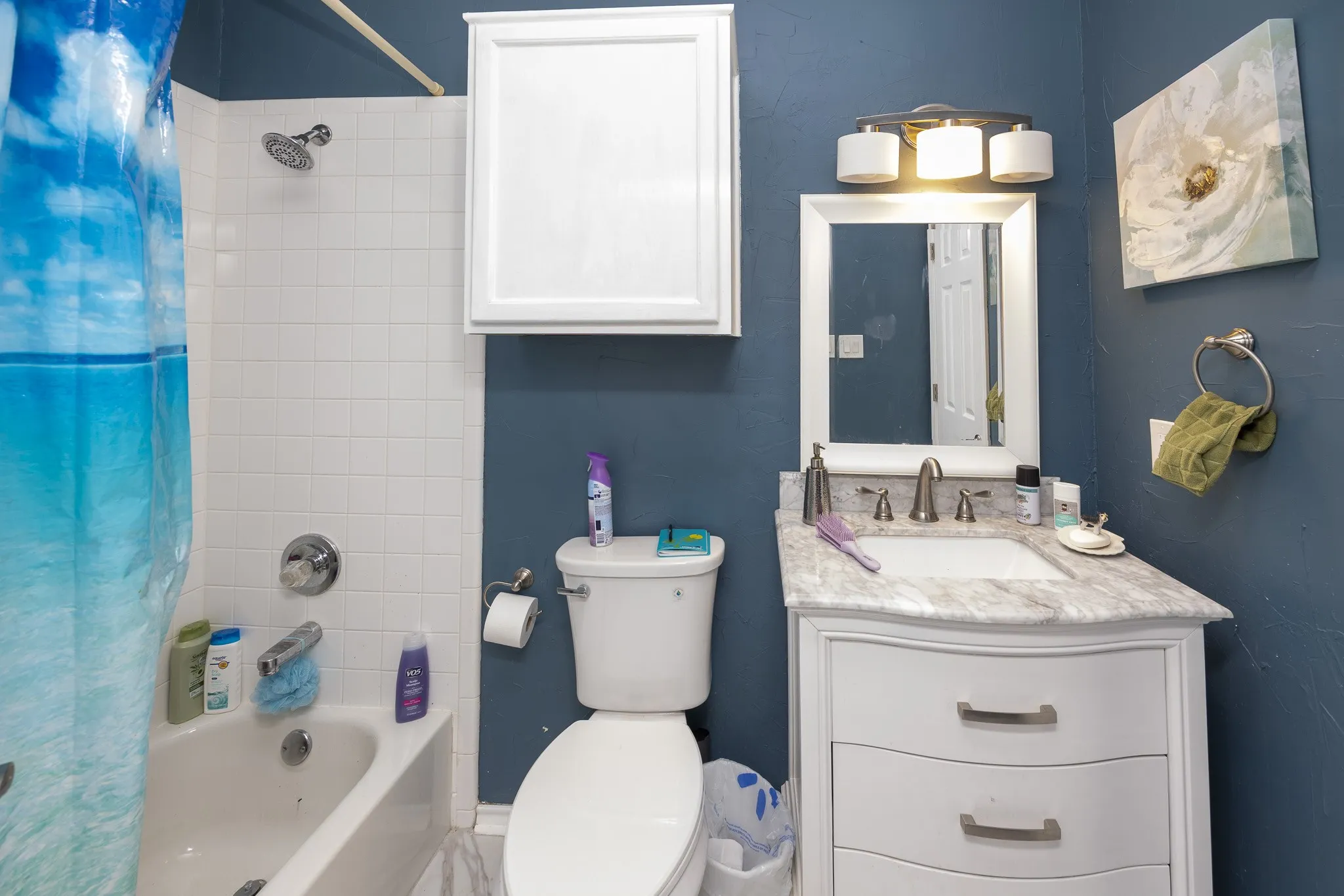 White subway tile shower surround with chrome fixtures, white built-in cabinetry, vanity with undermount sink and countertop, and a wall-mounted mirror with overhead lighting