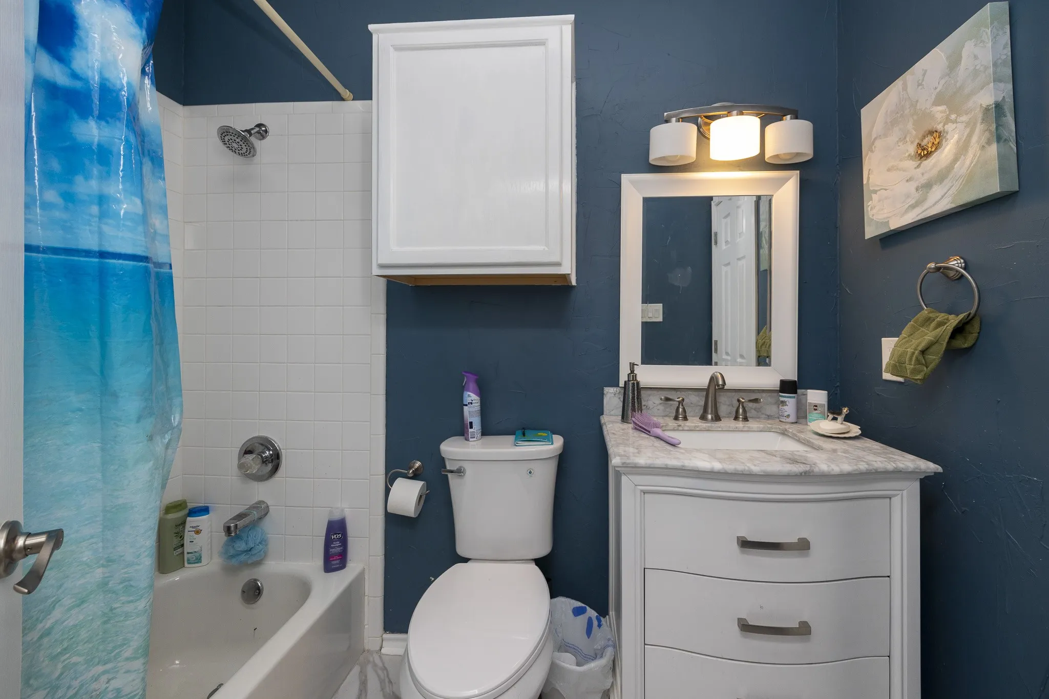 White vanity with a light-colored countertop, framed mirror, wall-mounted light fixture, white toilet, and white tile shower surround