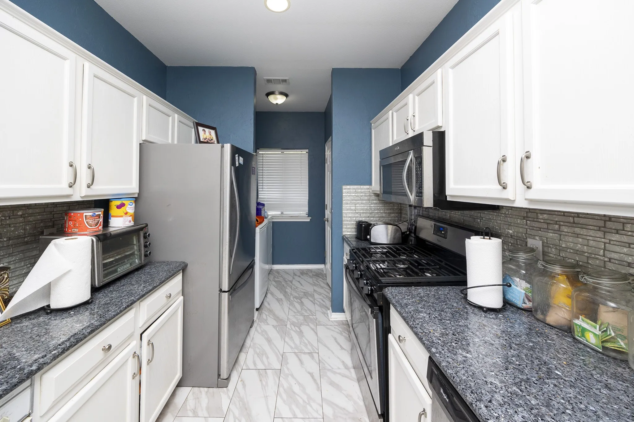 Galley kitchen featuring white cabinetry, dark speckled countertops, stainless steel appliances, a tiled backsplash, and light-toned flooring