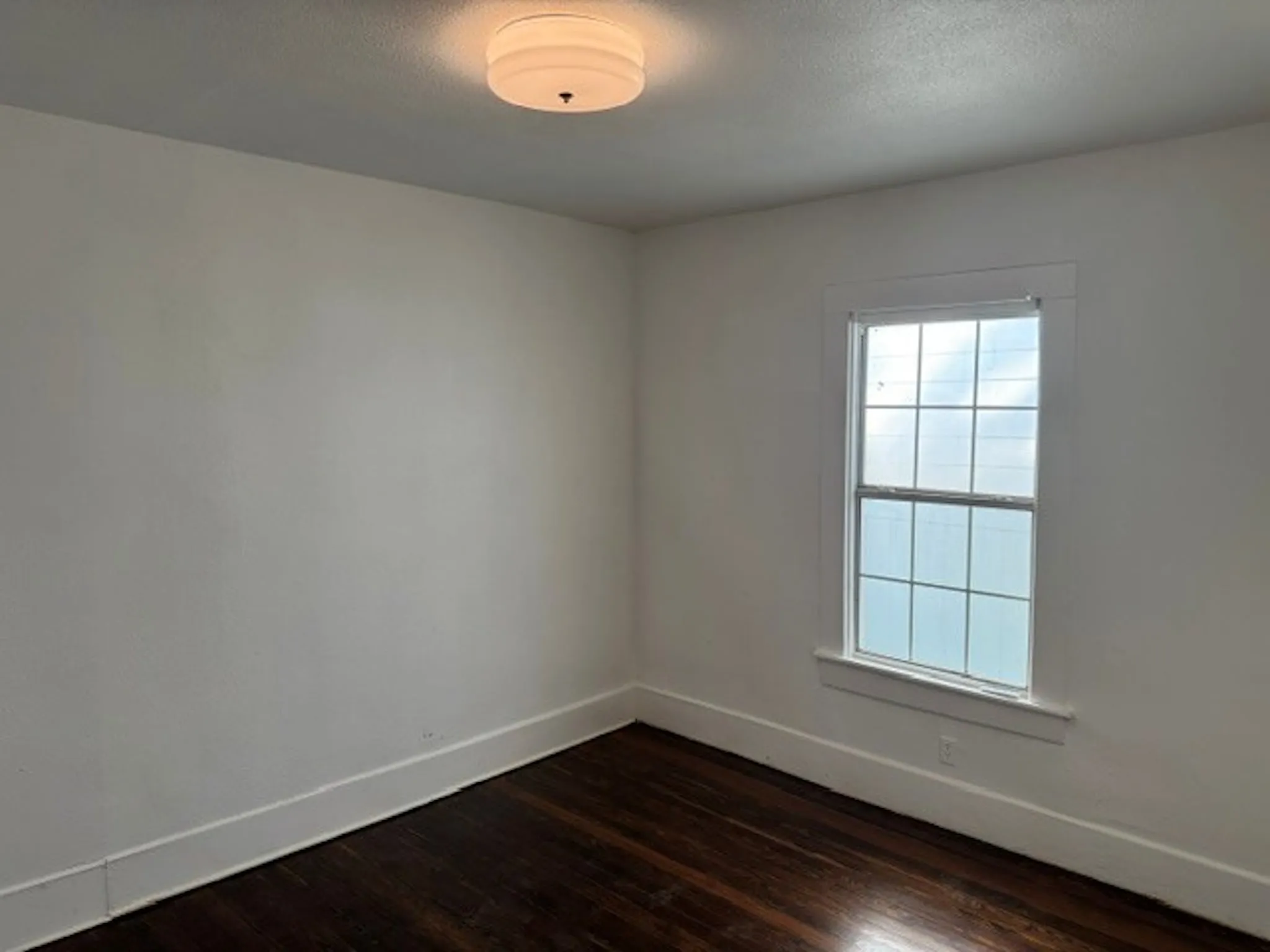 White walls with dark wood-finish flooring, featuring a single-pane window with white trim and a flush-mount ceiling light