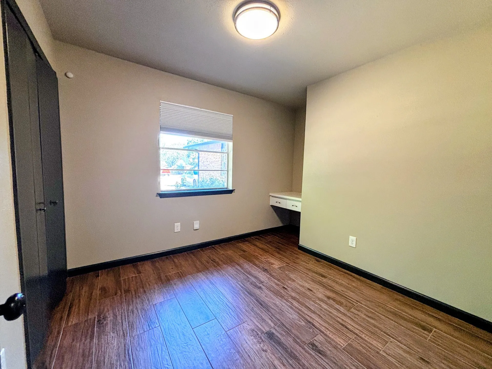 Wood-finish flooring, built-in desk, window with blinds, overhead flush mount lighting, and built-in closet with dark doors
