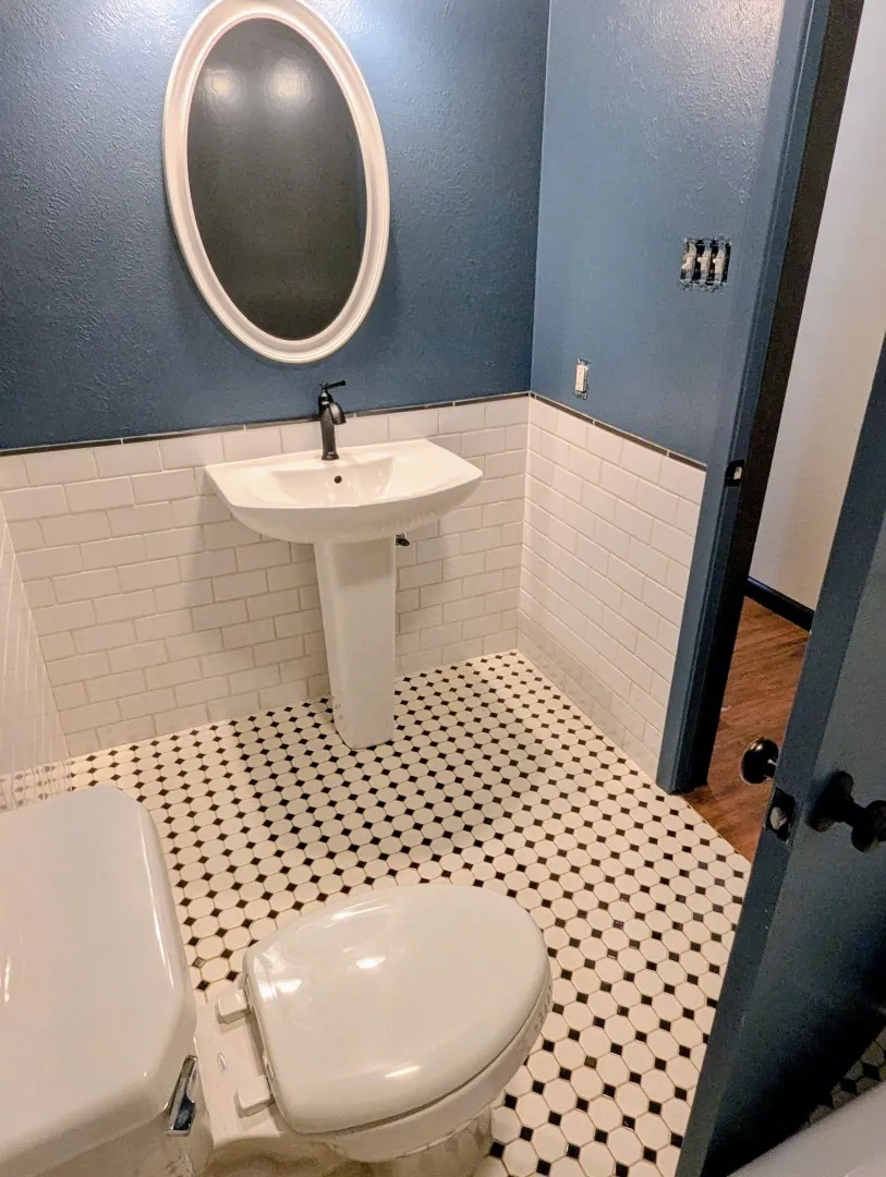 Pedestal sink with a matte black faucet, wall-mounted oval mirror, white subway tile wainscoting, and black and white octagon tile flooring