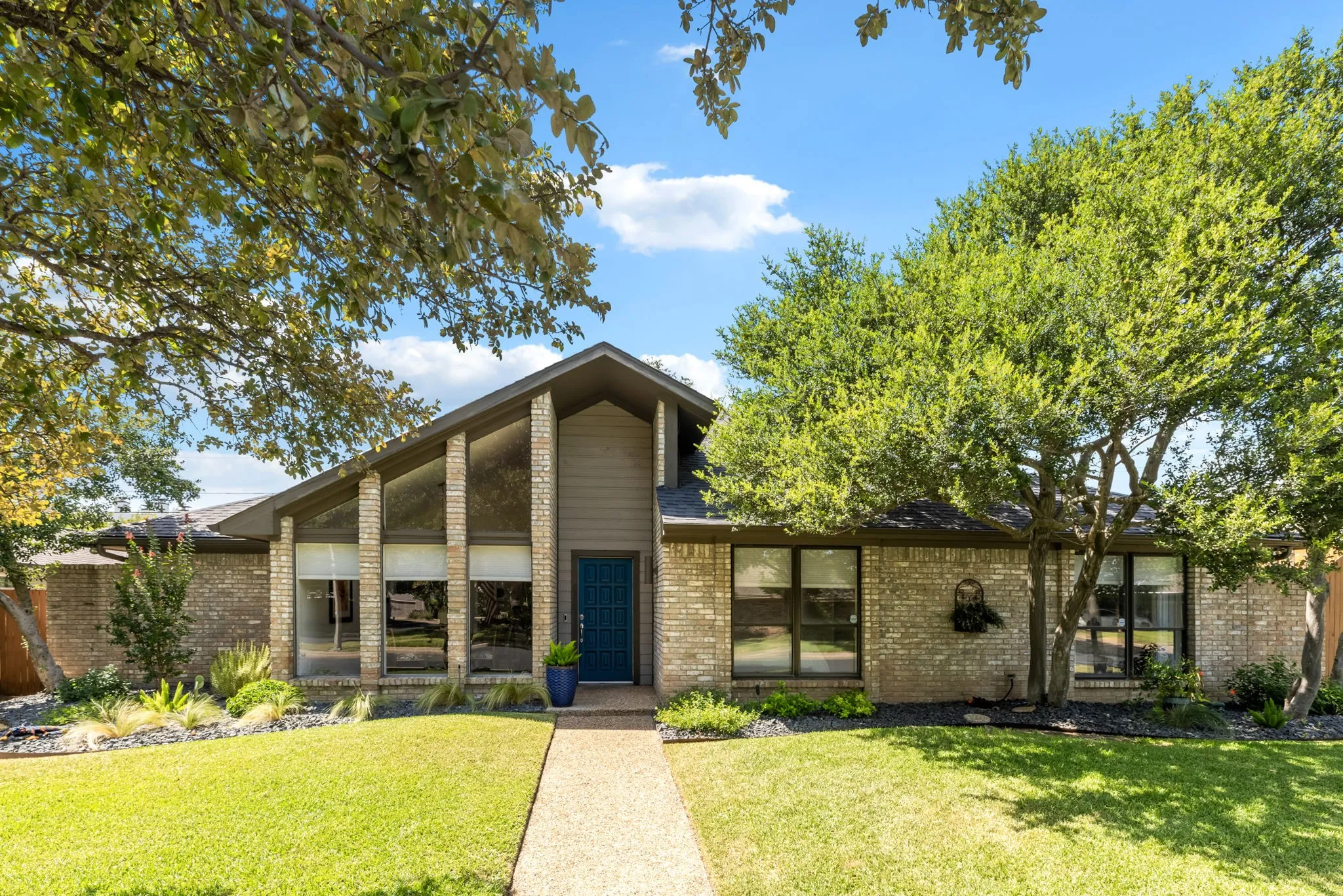 Brick exterior with a peaked roofline, a striking blue entry door, expansive front windows, mature trees, and a concrete walkway