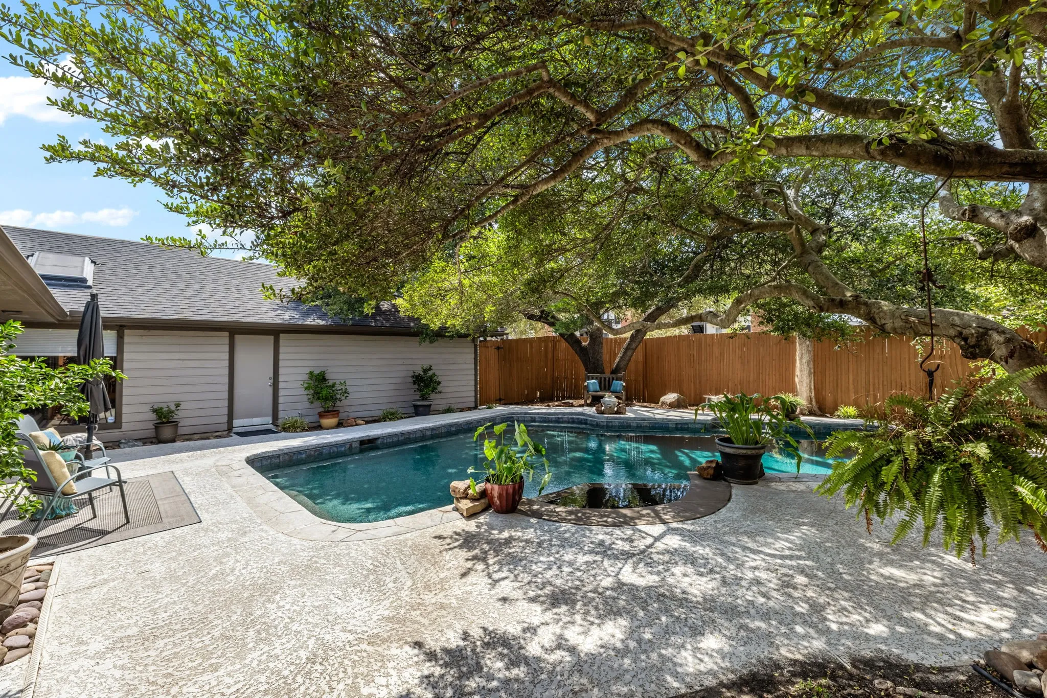 Private backyard featuring a swimming pool with a waterfall, mature trees, concrete patio, and a wood privacy fence