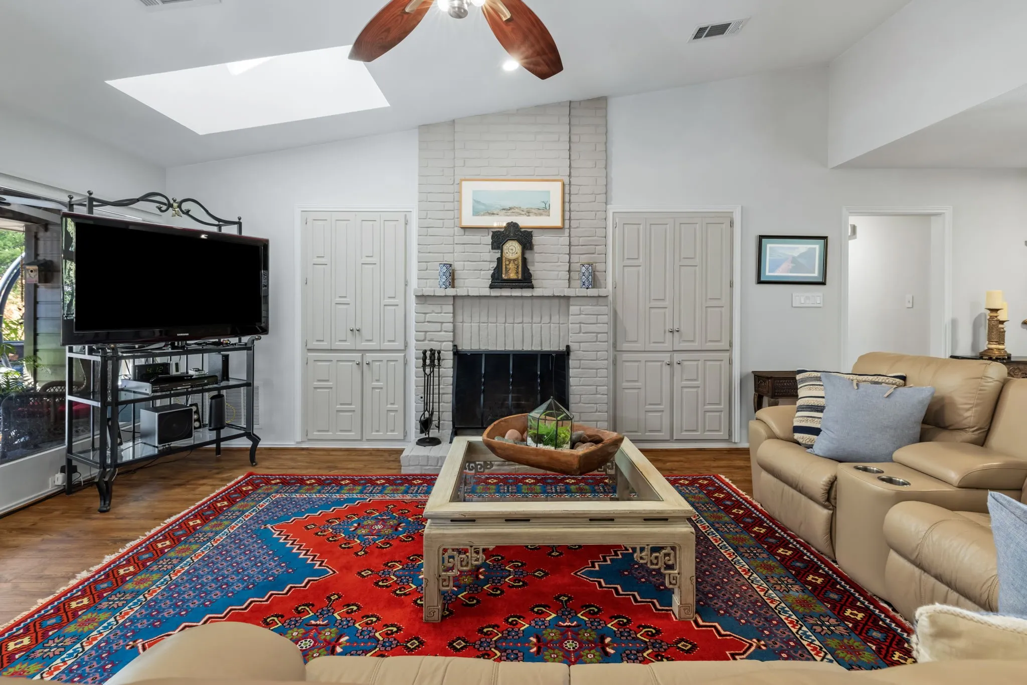 Vaulted ceiling with skylight, brick fireplace, wood-finish flooring, ceiling fan, and built-in cabinetry