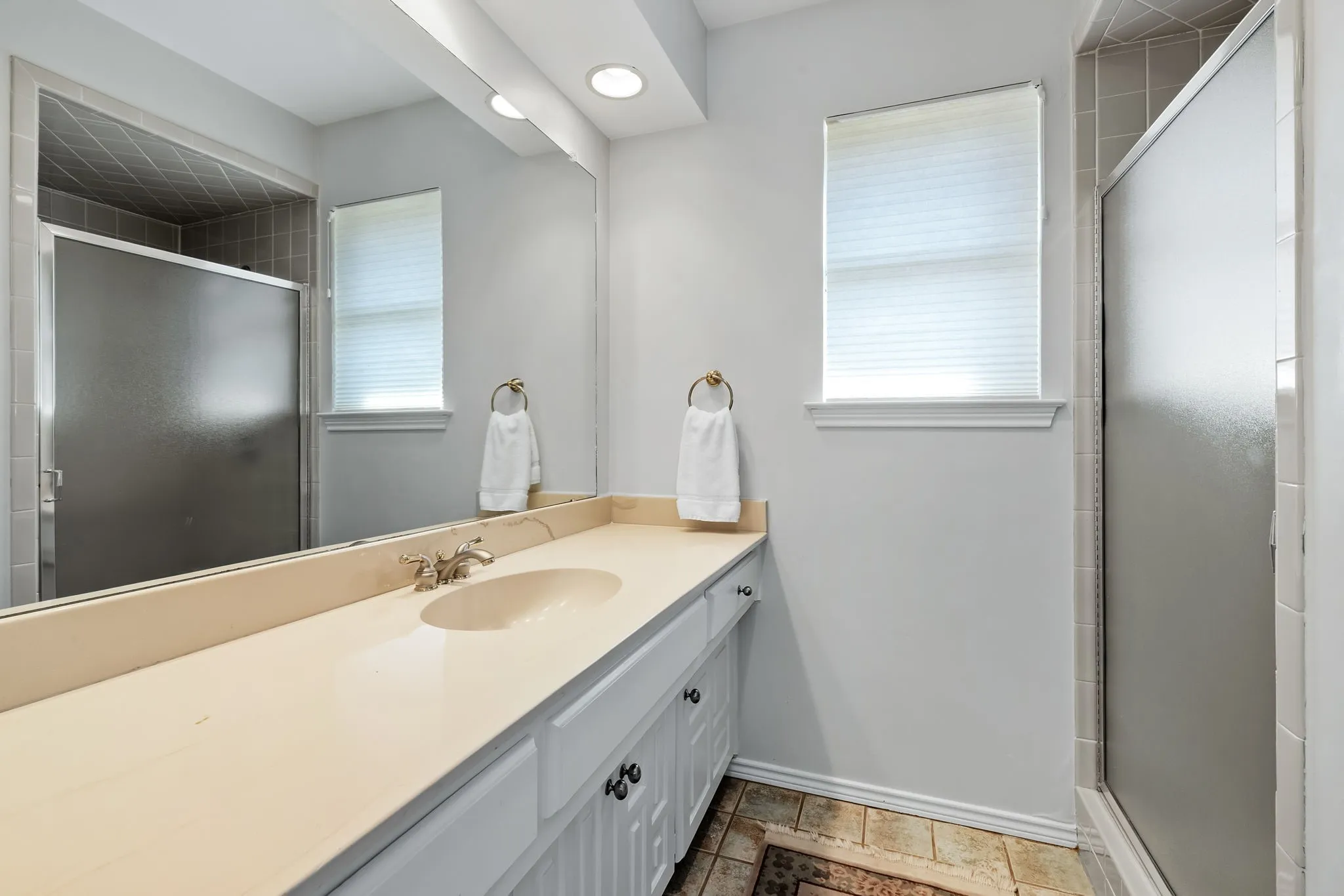 Dual vanity with an integrated sink and brushed nickel faucet, large wall mirror, recessed lighting, and a walk-in shower with frosted glass doors