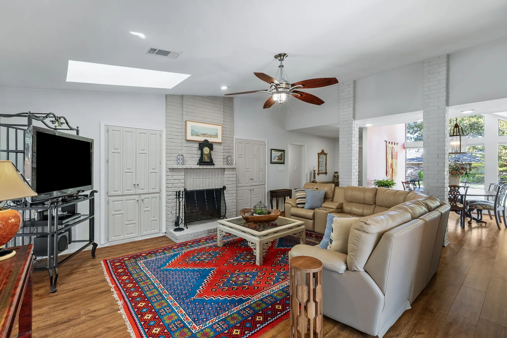 Vaulted ceiling with skylight, wood-finish flooring, white-painted brick fireplace, built-in cabinetry, and ceiling fan with lighting