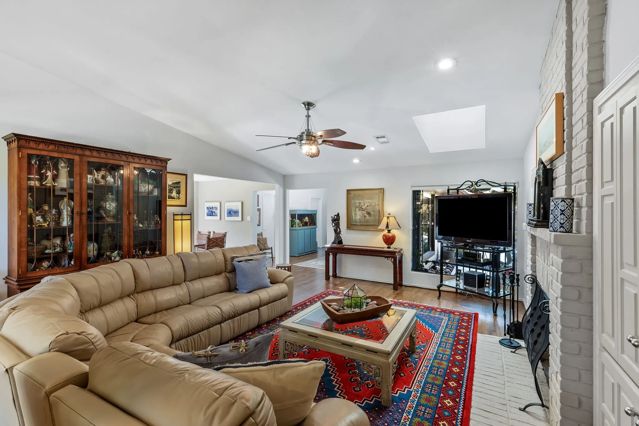 Vaulted ceiling with a ceiling fan, recessed lighting, wood-finish flooring, white brick fireplace with mantel, and a skylight