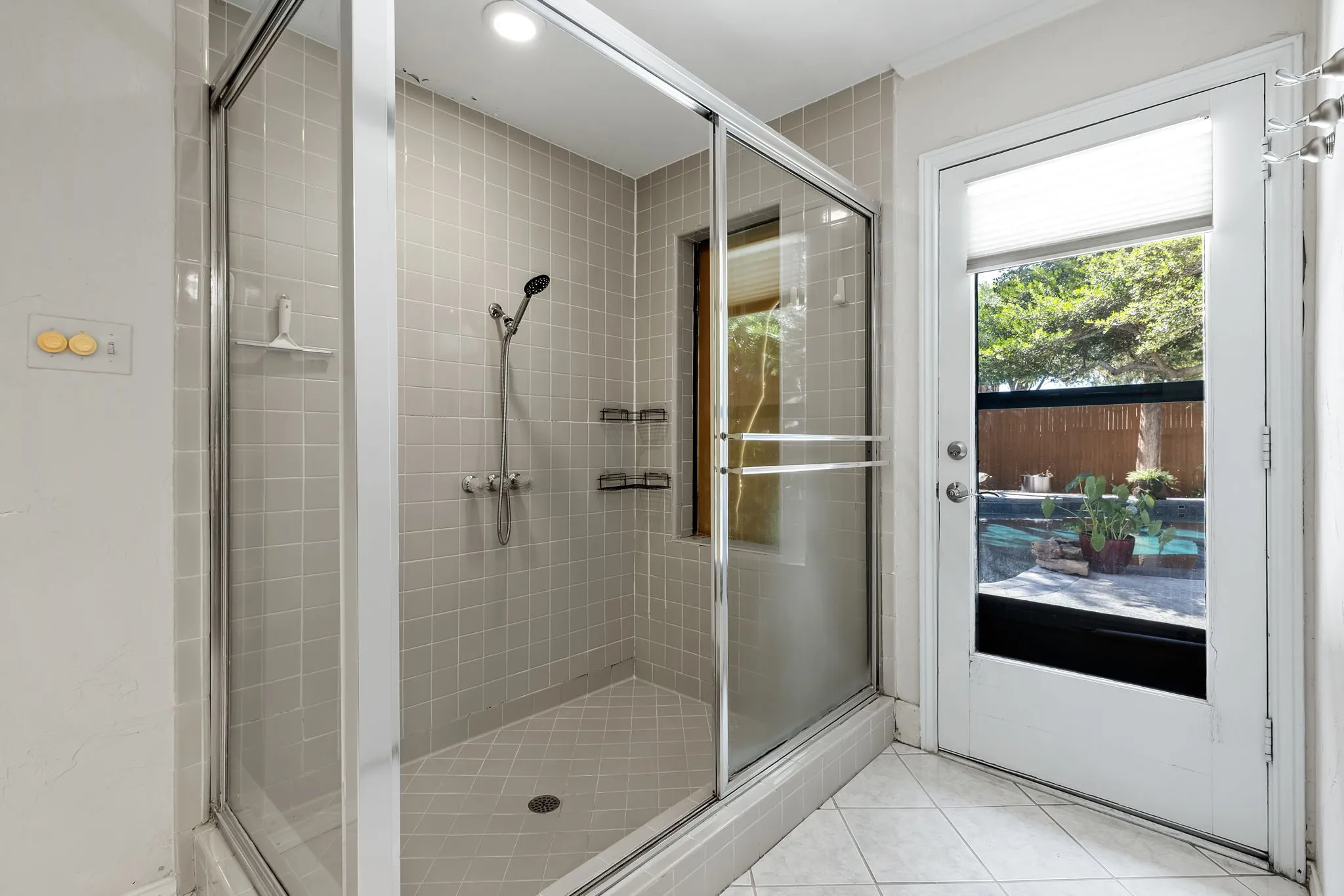 Tiled shower enclosure with a glass sliding door, built-in shelving, and an adjacent exterior door with a privacy blind
