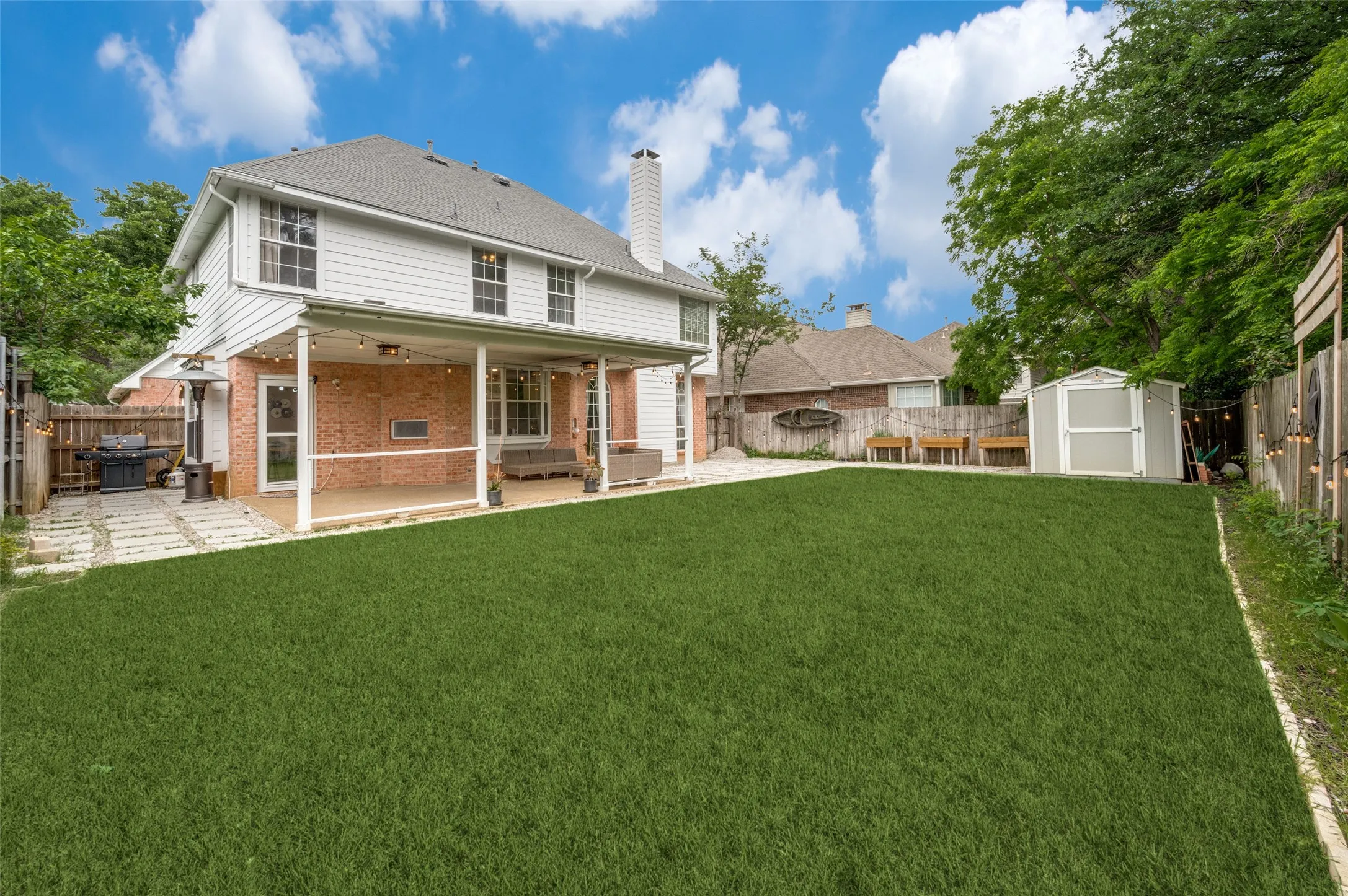Expansive green lawn with a covered patio featuring string lighting, a brick exterior, and a detached storage shed