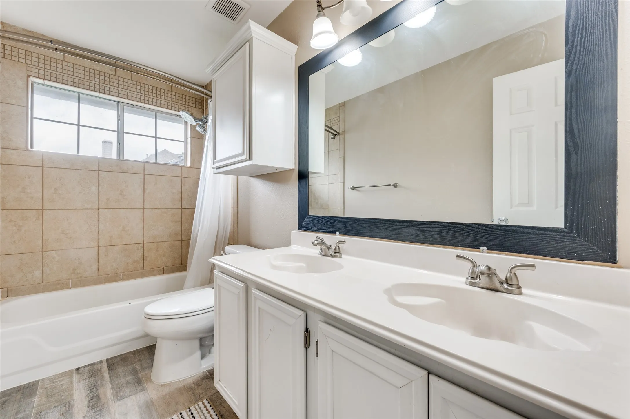Dual sink vanity with white cabinetry and brushed nickel fixtures, subway tile tub surround, window with horizontal blinds, and wood-finish flooring
