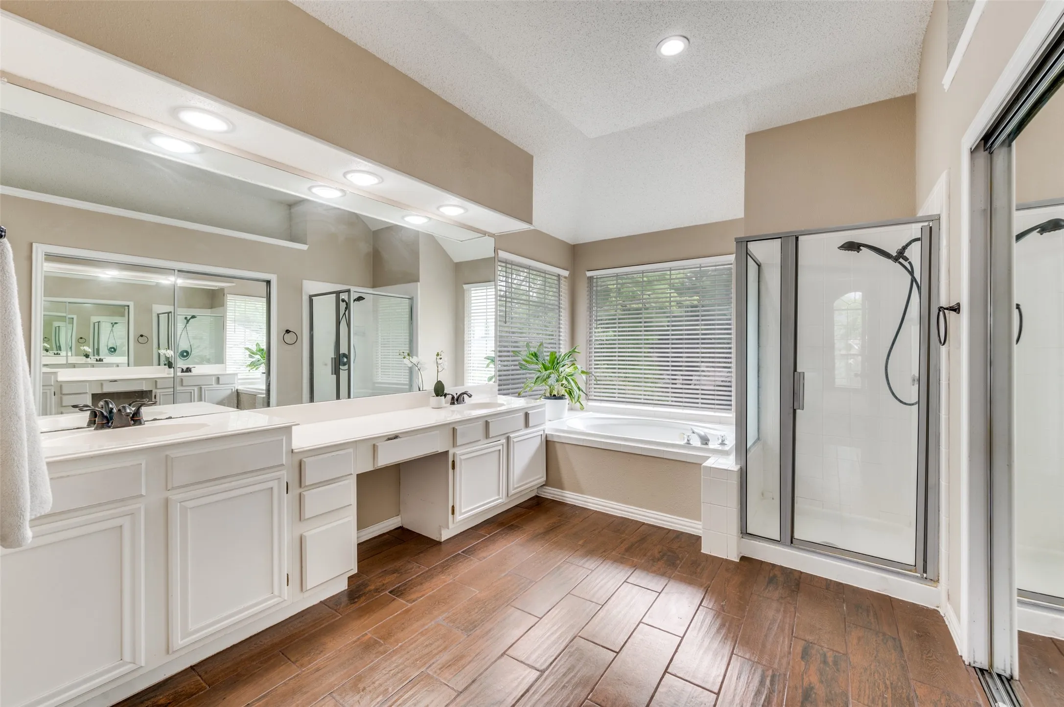 Spa-inspired primary bathroom showcasing dual vanities with white cabinetry, an expansive quartz countertop, recessed lighting, a soaking tub positioned at the window, a walk-in shower with glass enclosure and brushed nickel fixtures, and warm wood-finish flooring throughout