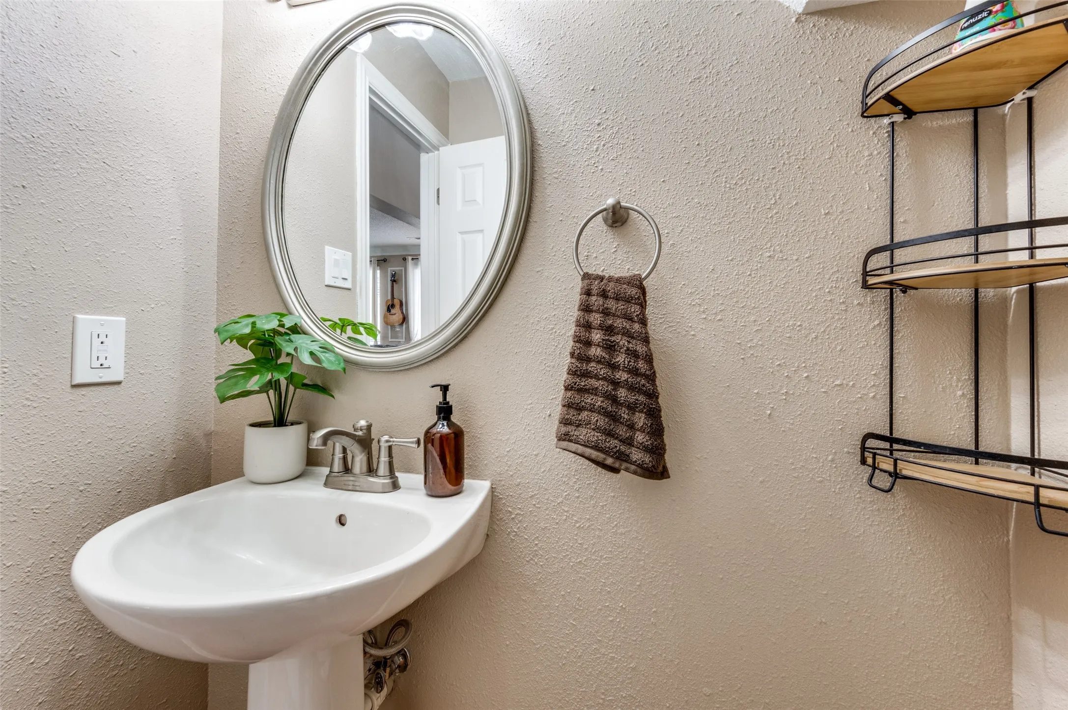 Pedestal sink with a brushed nickel faucet, oval framed mirror, and wall-mounted shelving