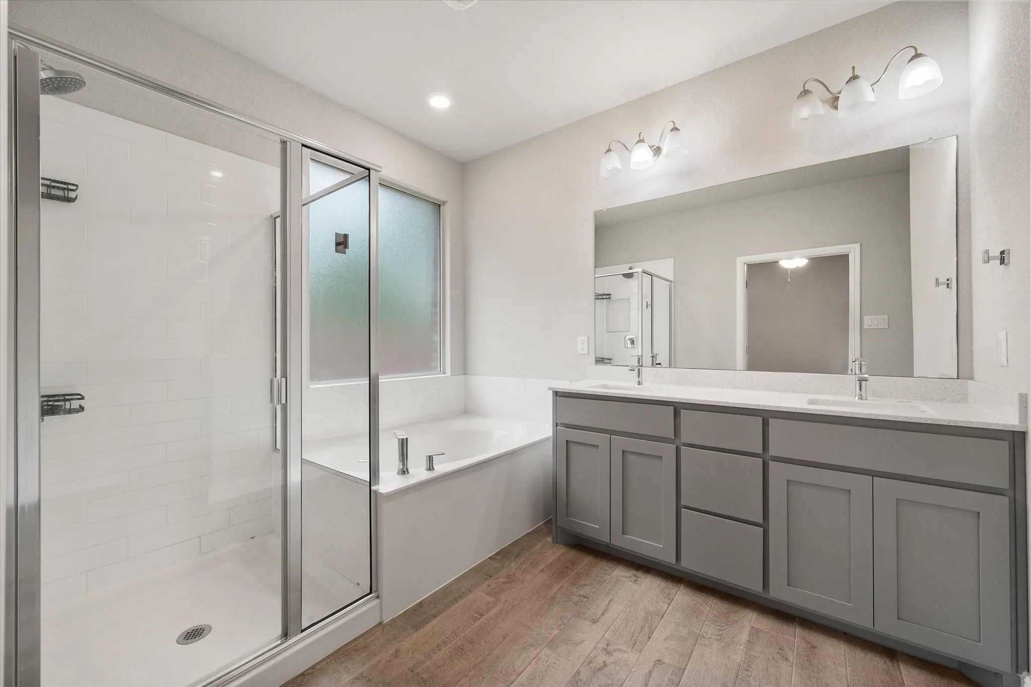 Spacious bathroom featuring a glass shower enclosure with subway tile surround, a built-in soaking tub, a dual vanity with gray shaker cabinetry and light-toned countertops, and wood-look tile flooring. There is a nice-sized walk-in closet off for the primary suite too!