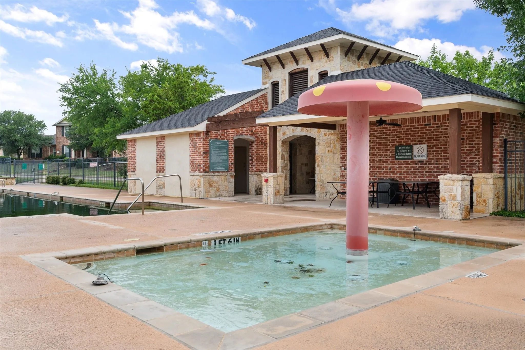 Spacious community pool area featuring a brick and stone building with a covered patio, a shallow water play area with a mushroom-shaped water feature, and a separate pool with handrails
