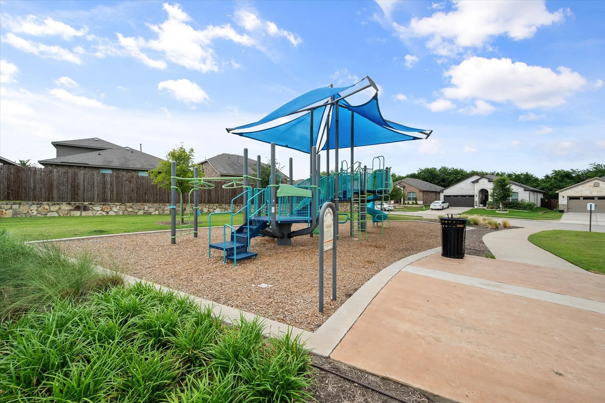 Community playground with shade canopy, adjacent to paved pathways, surrounded by green landscaping and individual residences