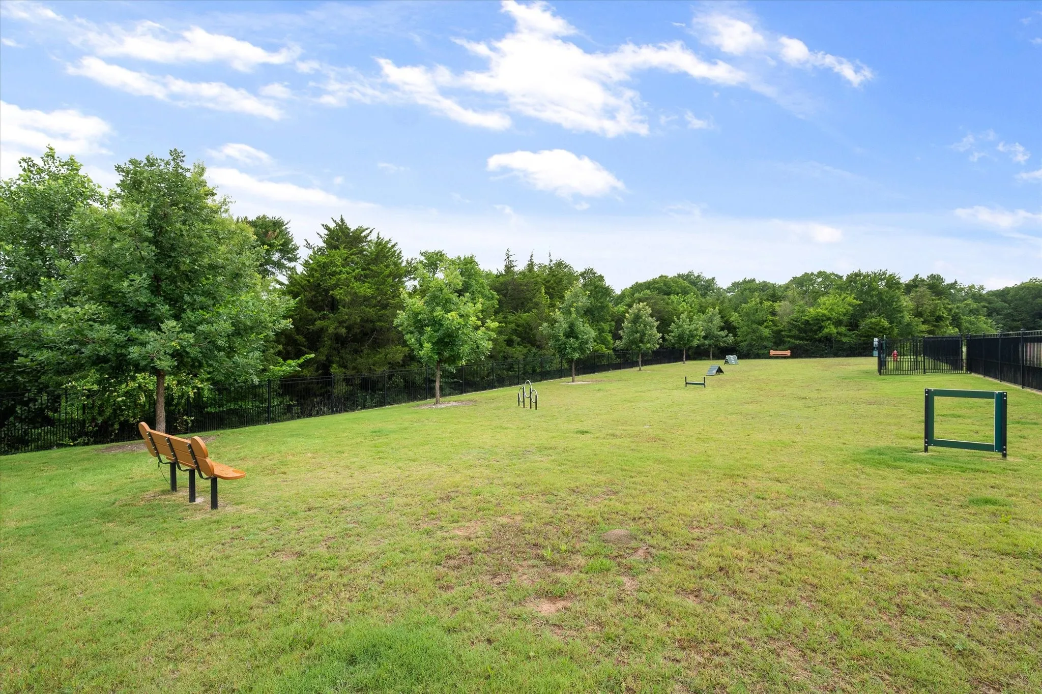 Dog Park has an expansive grassy area featuring a wooden bench, mature trees, and fenced perimeter