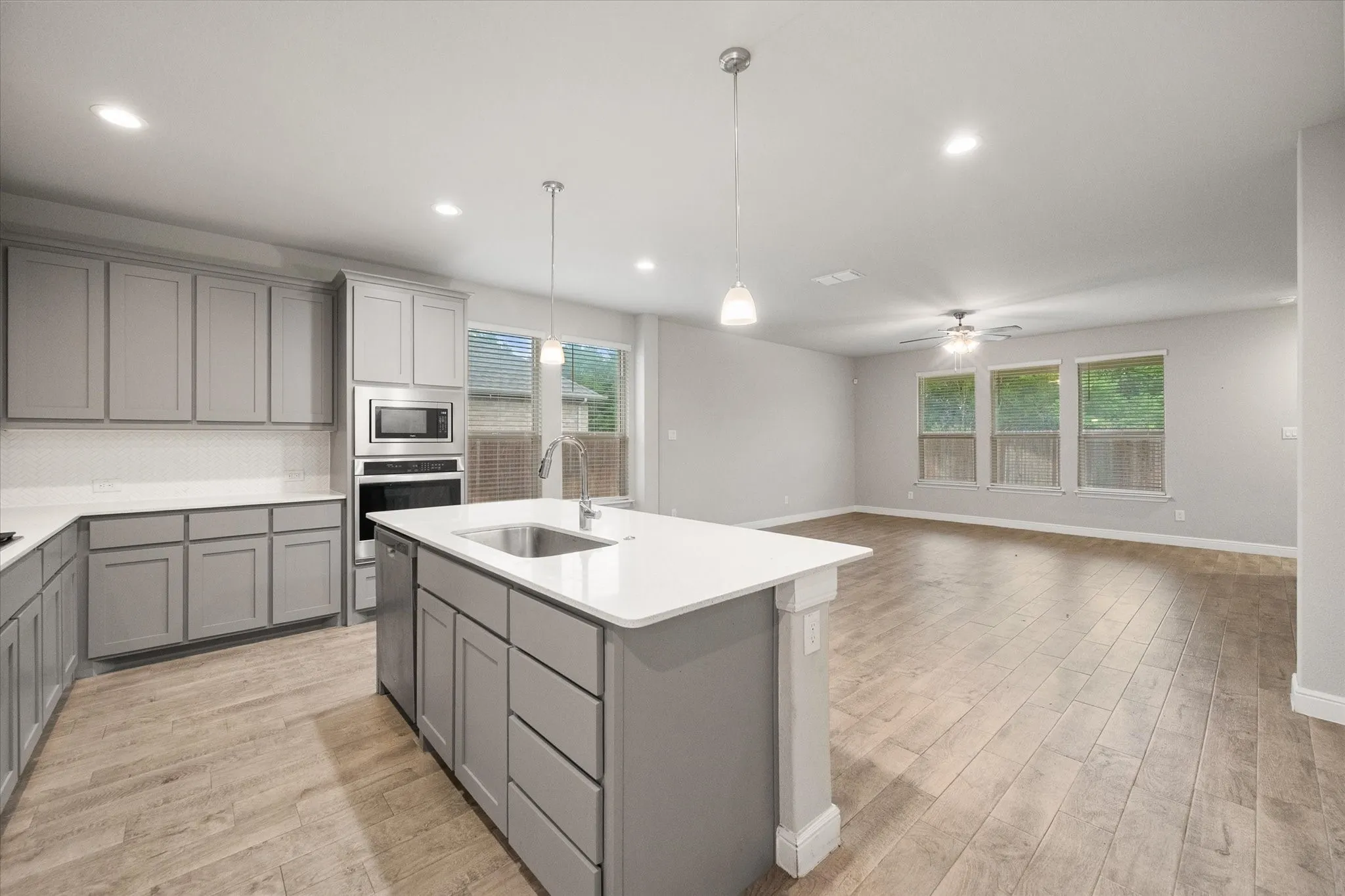 Open-concept kitchen featuring an island with an undermount sink, gray cabinetry, built-in stainless steel appliances, and wood-look tile flooring extending into the living & dining areas
