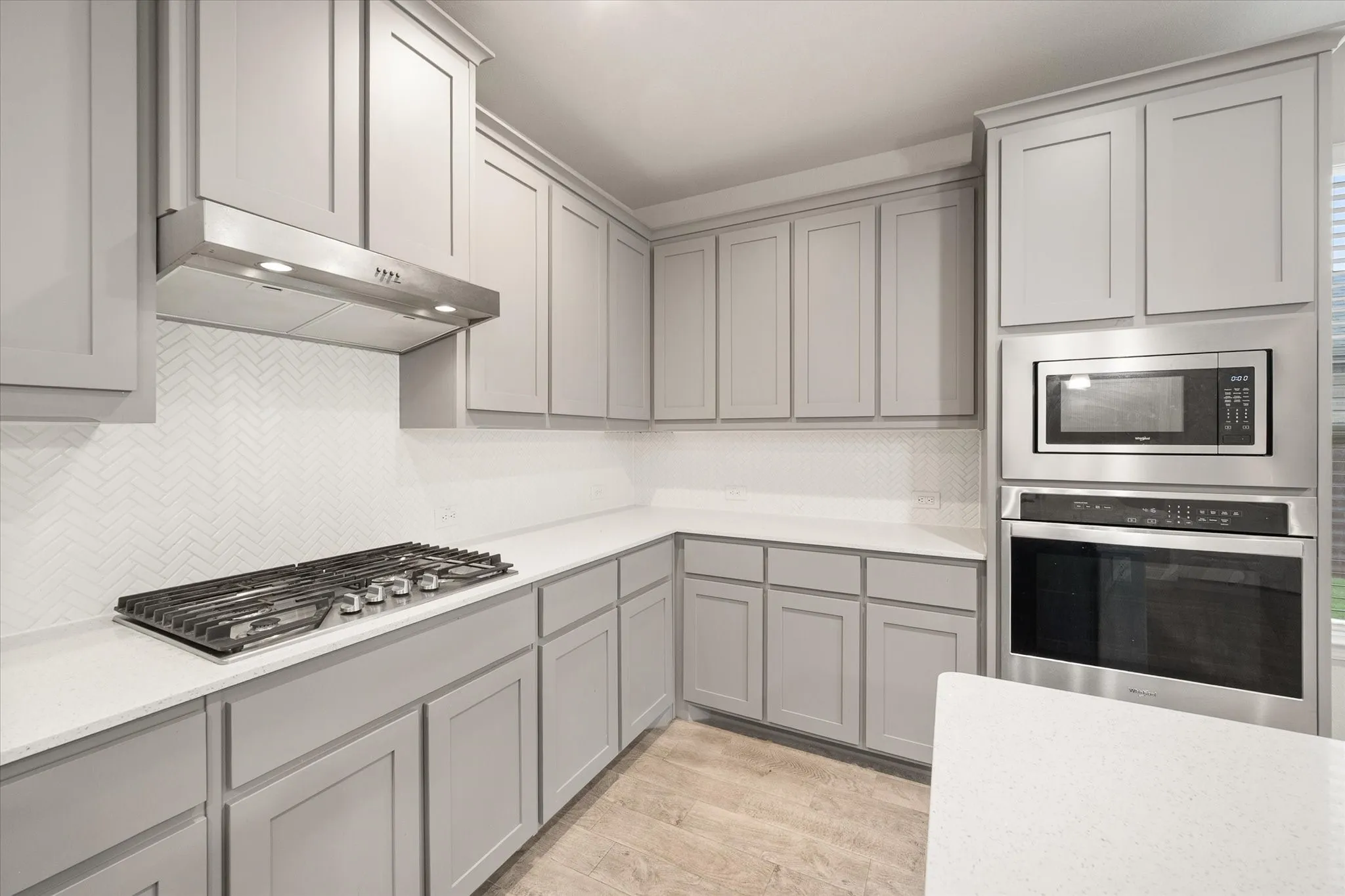 Light gray cabinetry, quartz countertops, herringbone tile backsplash, stainless steel gas cooktop, and built-in stainless steel microwave and oven