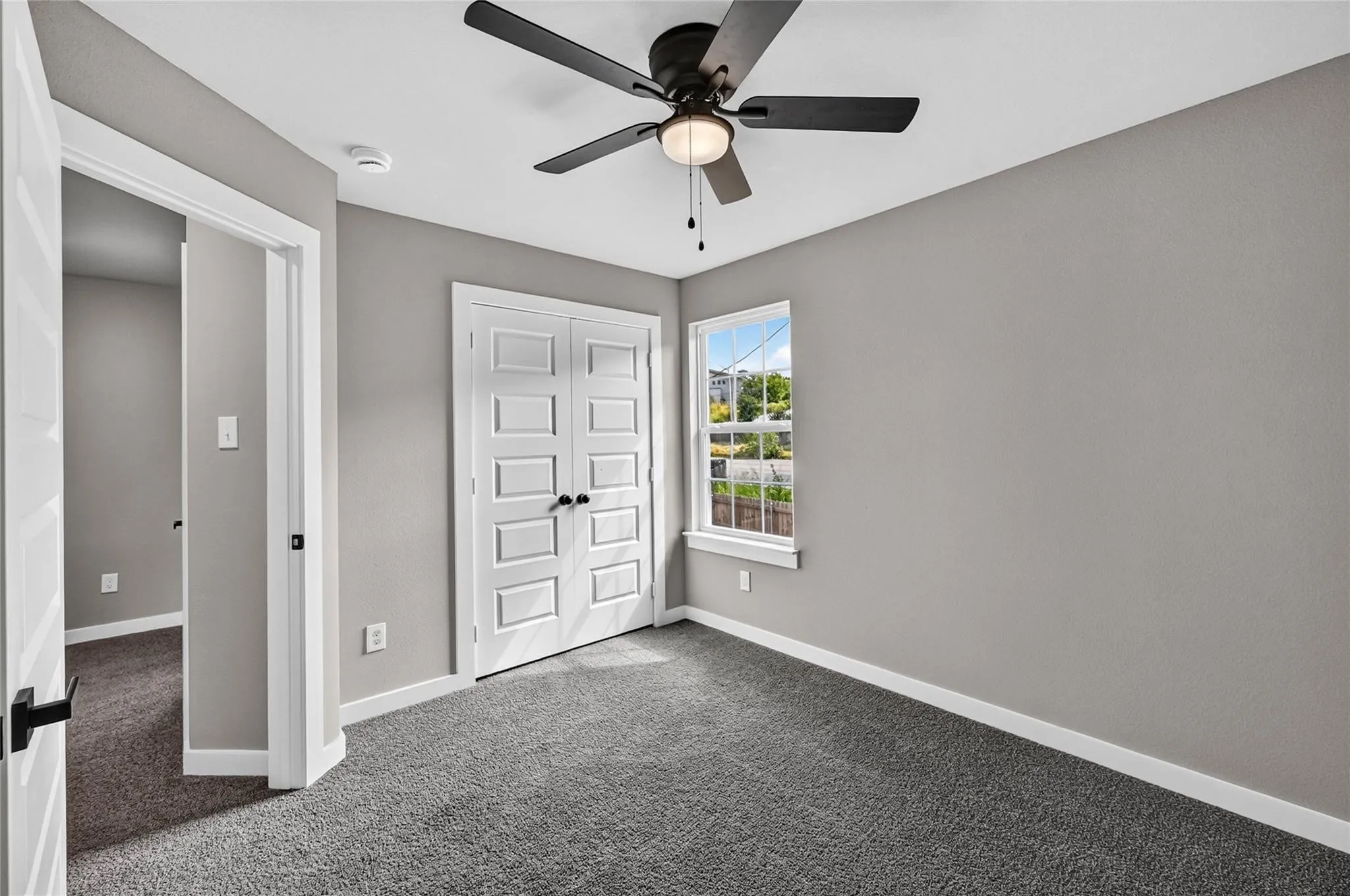 Ceiling fan with integrated lighting, window with white trim, gray textured walls, white paneled bifold closet doors, and gray carpeting