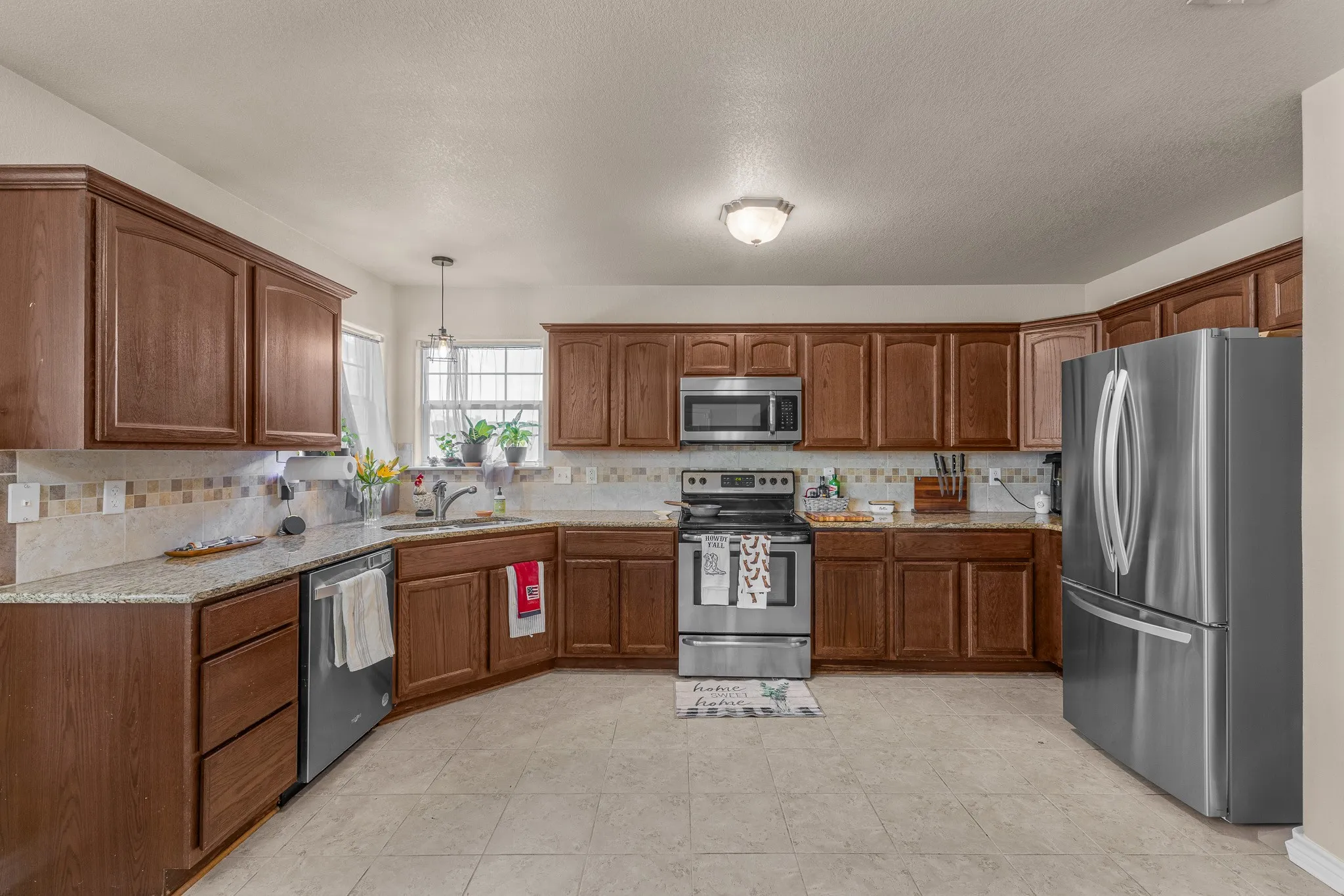 U-shaped kitchen featuring wood cabinetry, light-toned countertops, a multi-tone tile backsplash, stainless steel appliances, and tiled flooring