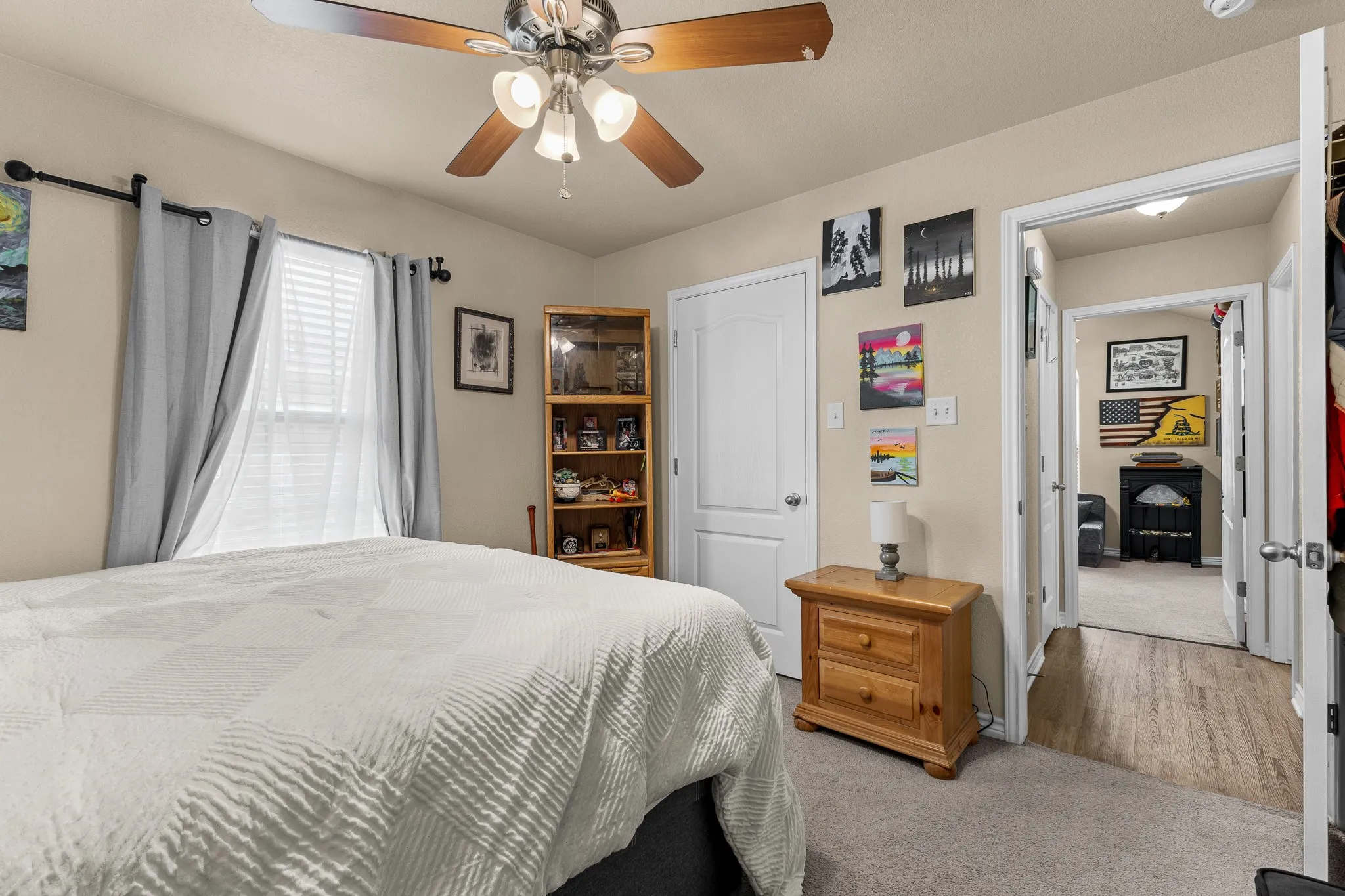 Ceiling fan with light fixture, window with blinds, carpeted flooring, wood-finish flooring in hallway, and a white interior door