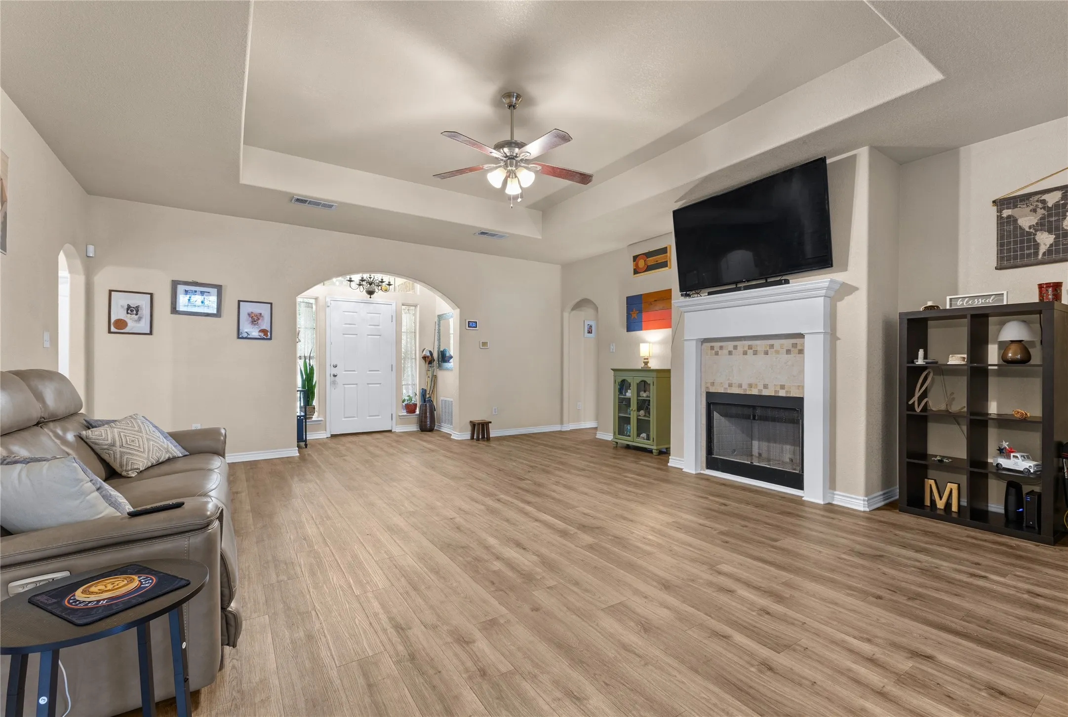 Tray ceiling with a ceiling fan, wood-finish flooring, a fireplace with a tiled surround and white mantel, and an arched entry