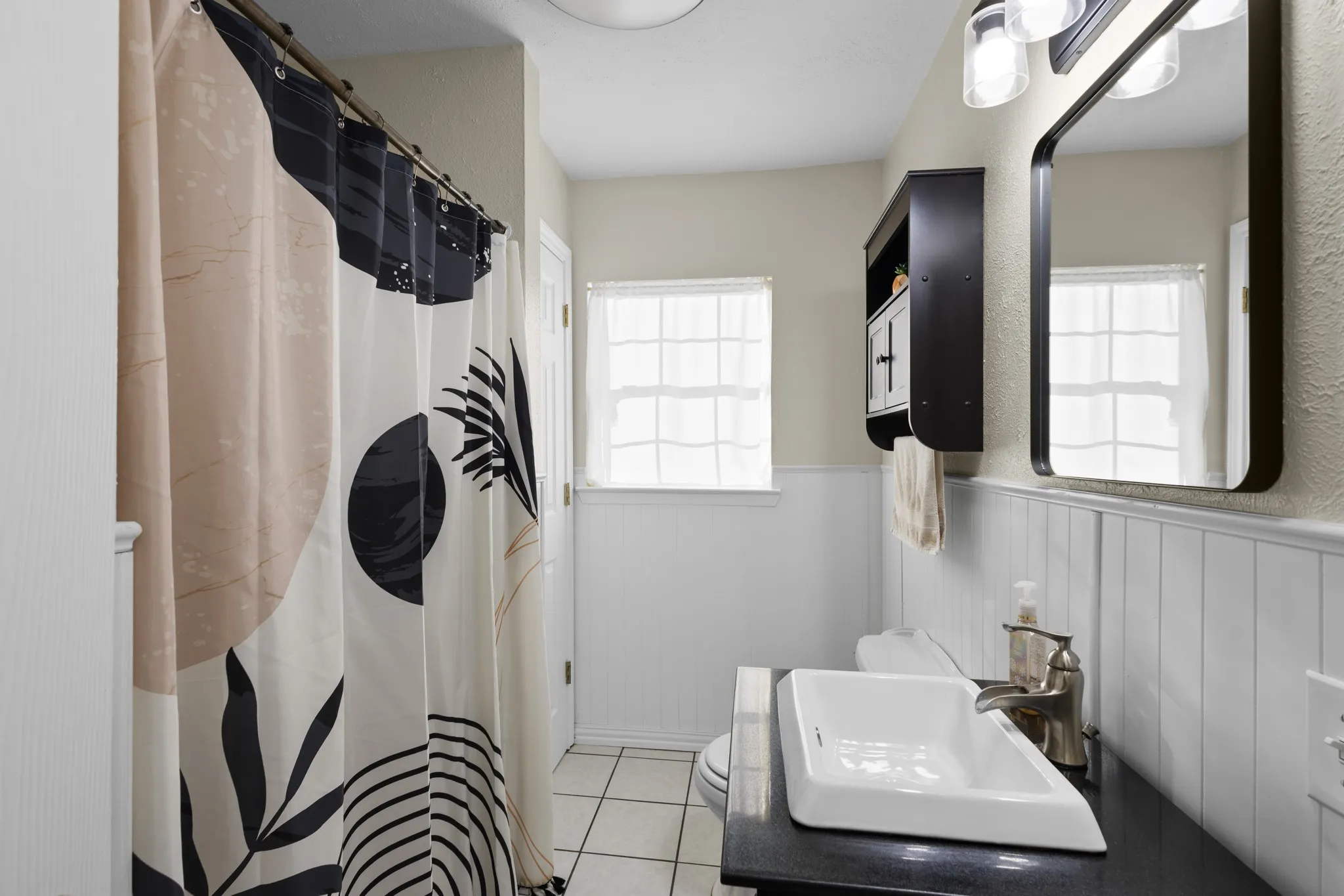 Bathroom featuring tile flooring, wainscoting, a window with natural light, a dark vanity countertop with an integrated sink, and a wall-mounted mirror with overhead lighting