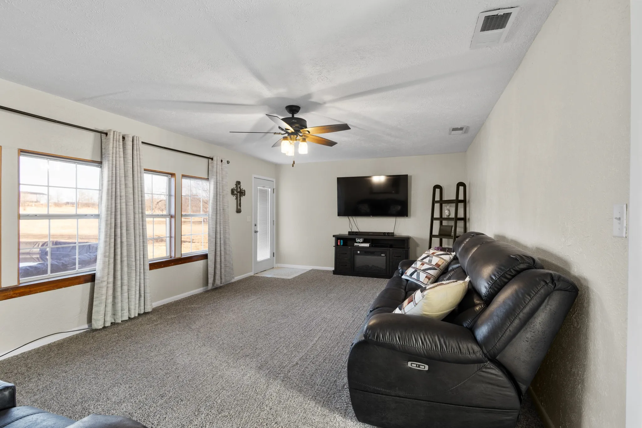 Carpeted flooring, multiple large windows, ceiling fan, neutral wall tones, and rear door with integrated glass panel