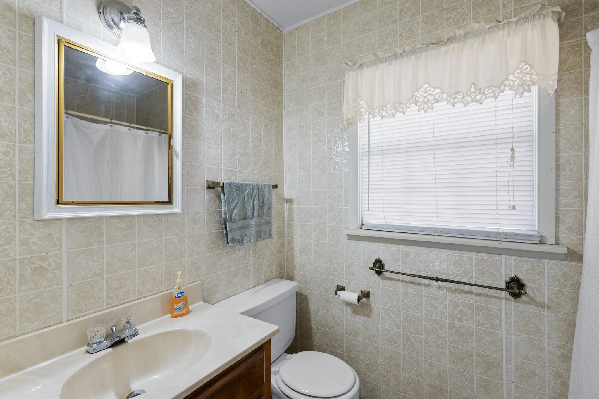 Tiled bathroom walls featuring a window with blinds, a vanity with an integrated sink, a toilet, and a medicine cabinet with an overhead light fixture